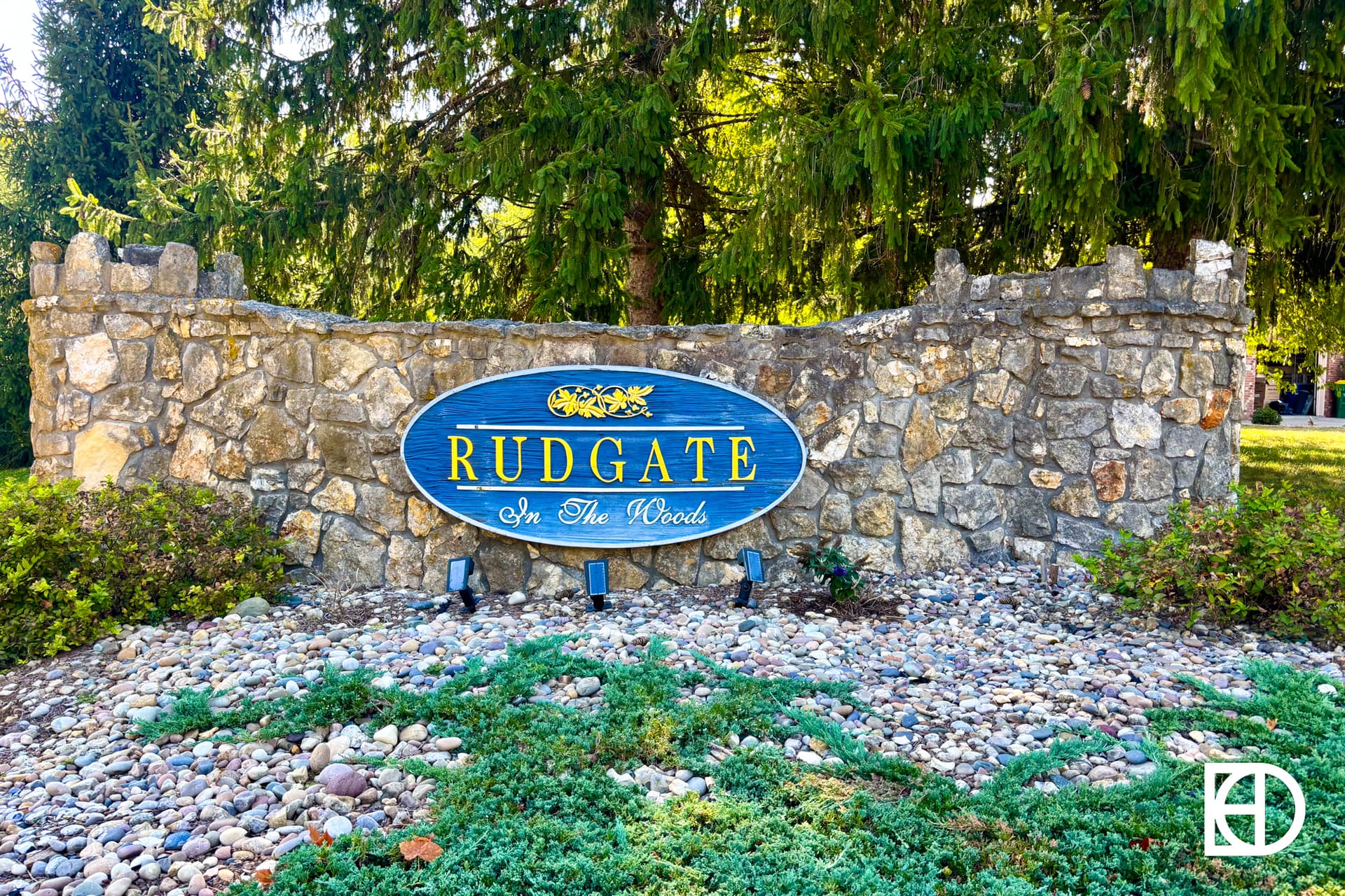 A stone wall with a blue oval sign reading Rudgate In The Woods in white and yellow letters, surrounded by rocks and greenery, with tall trees in the background.
