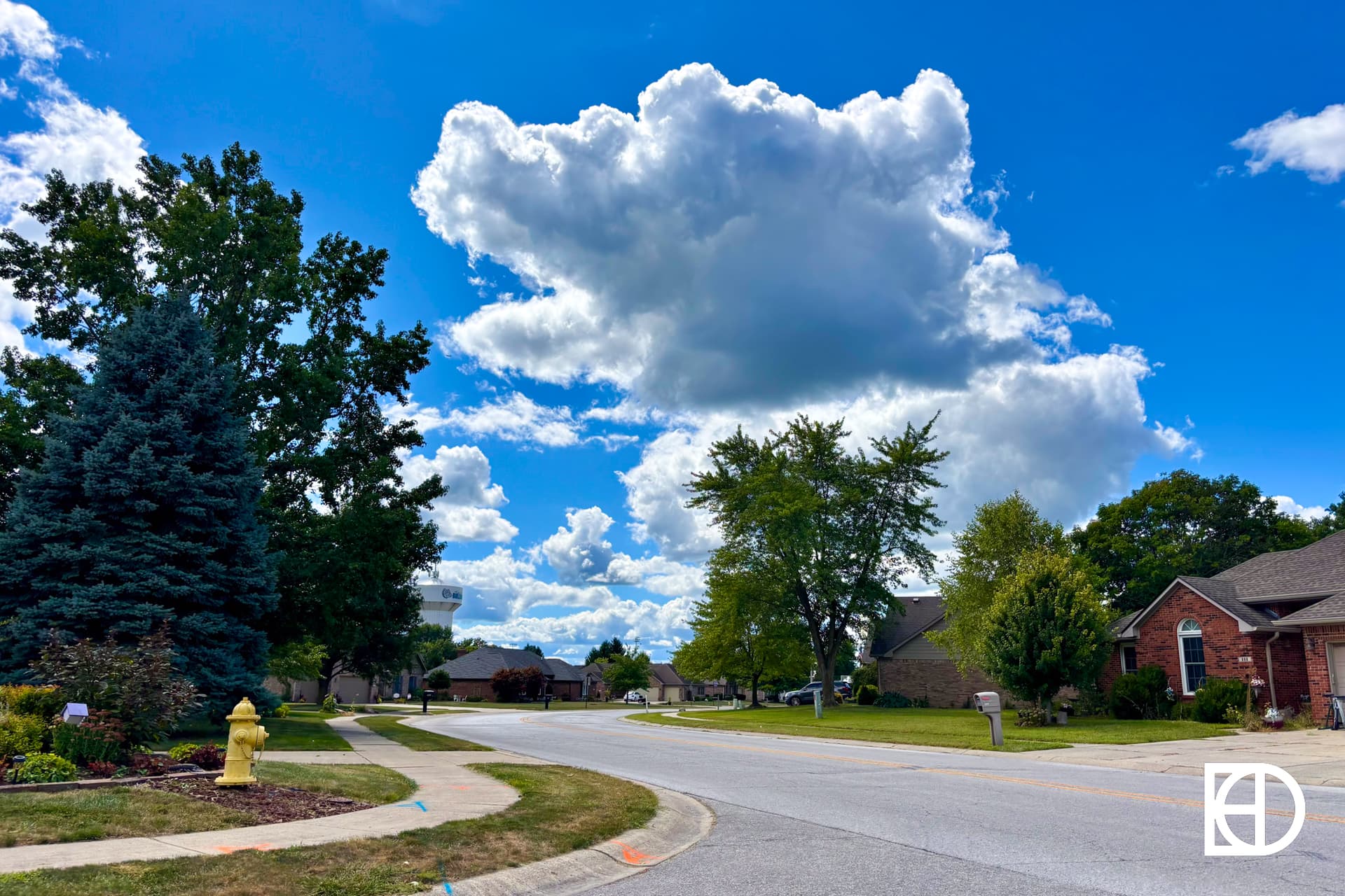 A suburban street on a sunny day with a bright blue sky and large, fluffy clouds. Trees and houses line the road, with a yellow fire hydrant and trimmed lawns in the foreground.