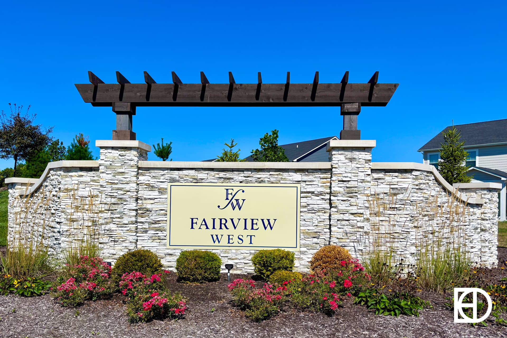 A stone entrance sign with a wooden pergola reads Fairview West in front of landscaped shrubs and flowers, set against a clear blue sky and nearby houses.