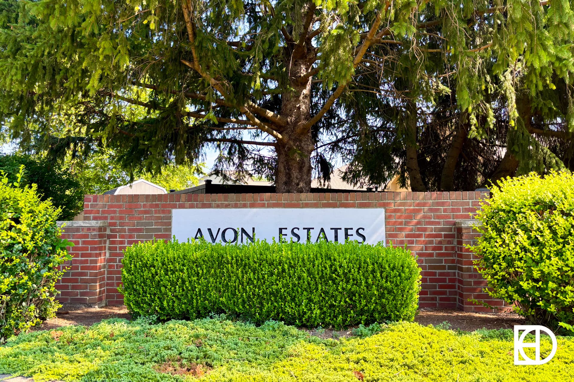 A brick sign reads AVON ESTATES surrounded by green hedges and trees, with sunlight filtering through the branches.