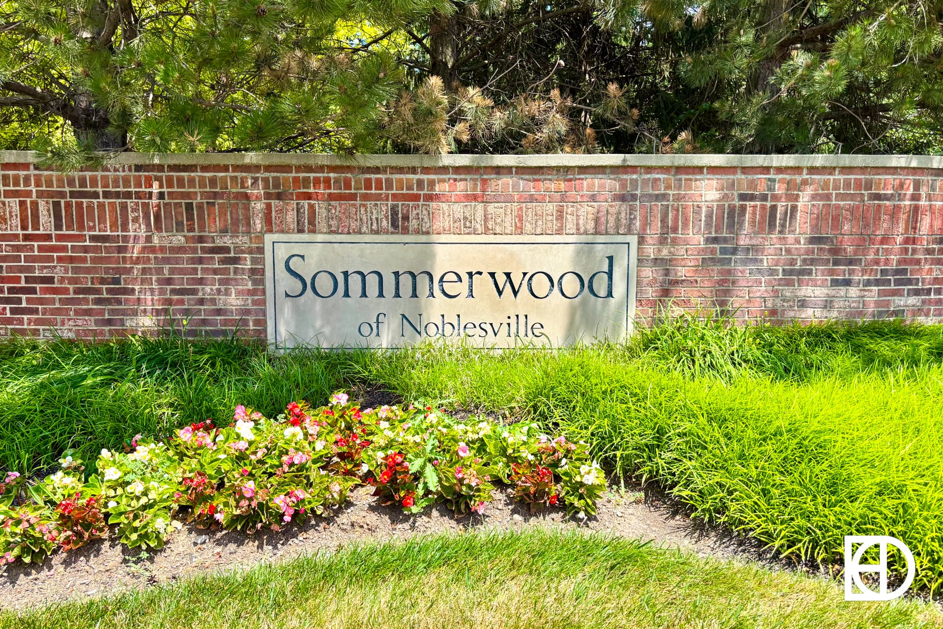 A brick sign reading Sommerwood of Noblesville stands behind a flower bed with red and white flowers and green grass, bordered by trees and shrubs.