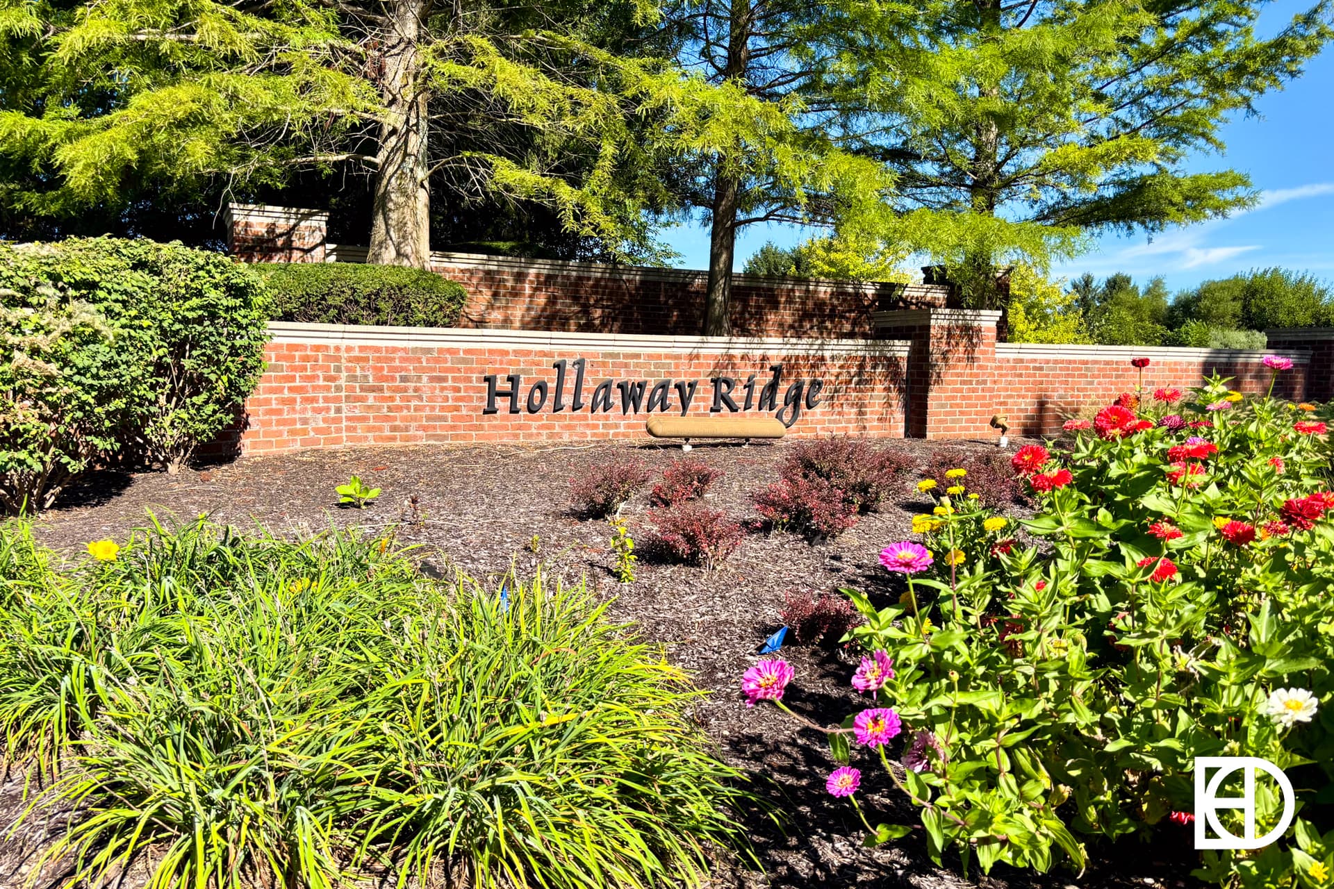 A brick sign reading Holloway Ridge is surrounded by green shrubs, colorful flowers, and trees under a bright blue sky.