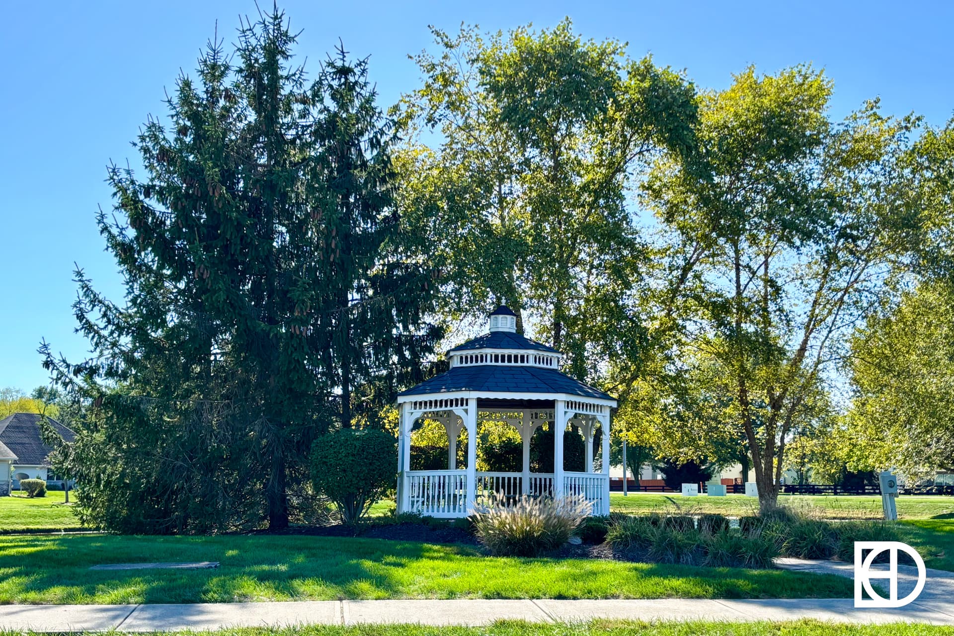 A white gazebo stands on a grassy lawn, partially shaded by large green trees under a clear blue sky. A sidewalk runs in front, and houses are visible in the background.