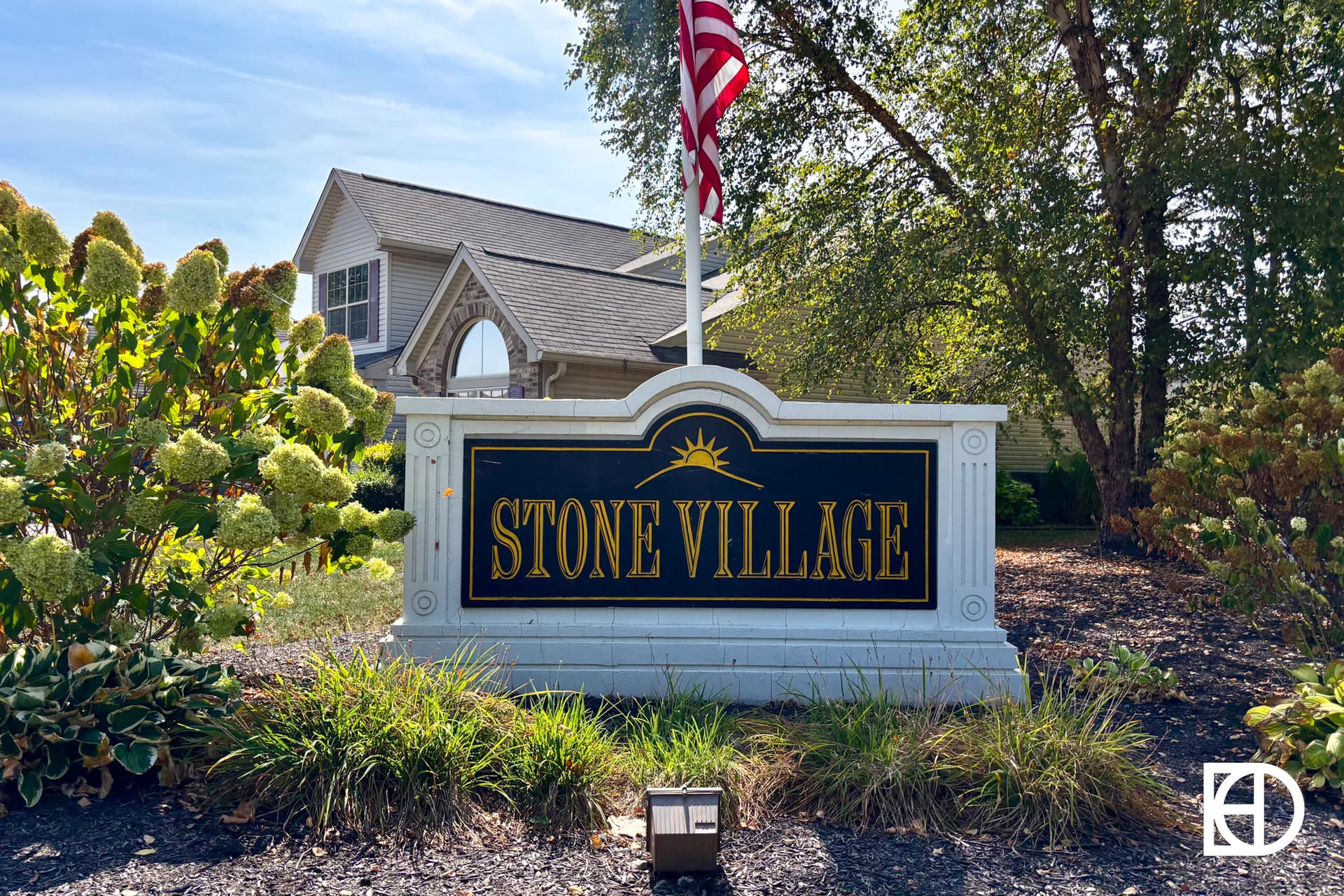 A sign reading Stone Village stands in front of a landscaped area with bushes and a tree, an American flag above it, and a house with large windows in the background.