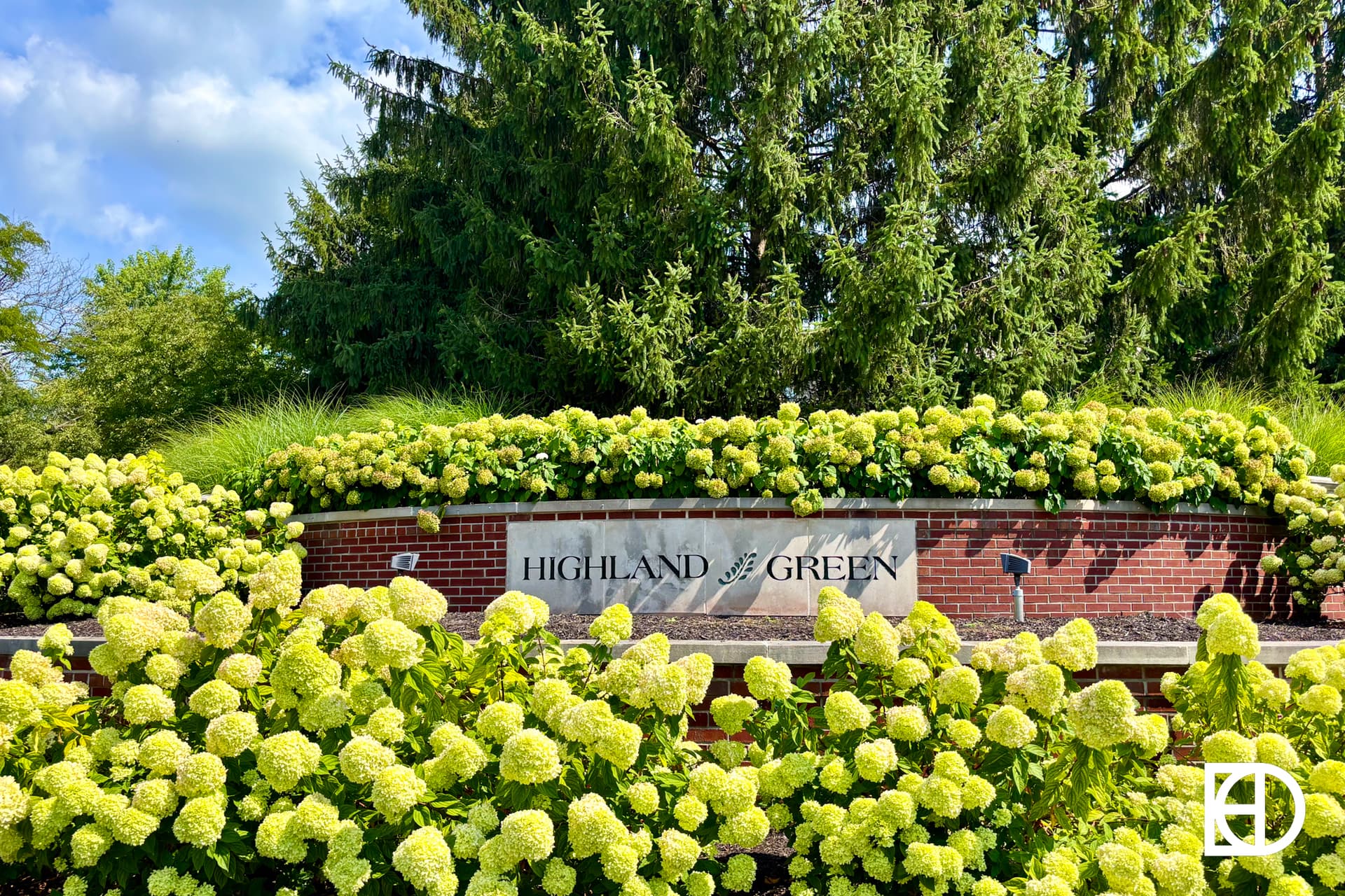 A landscaped brick sign surrounded by blooming hydrangeas reads HIGHLAND GREEN. Tall evergreen trees and a partly cloudy sky are in the background. A white logo is in the lower right corner.