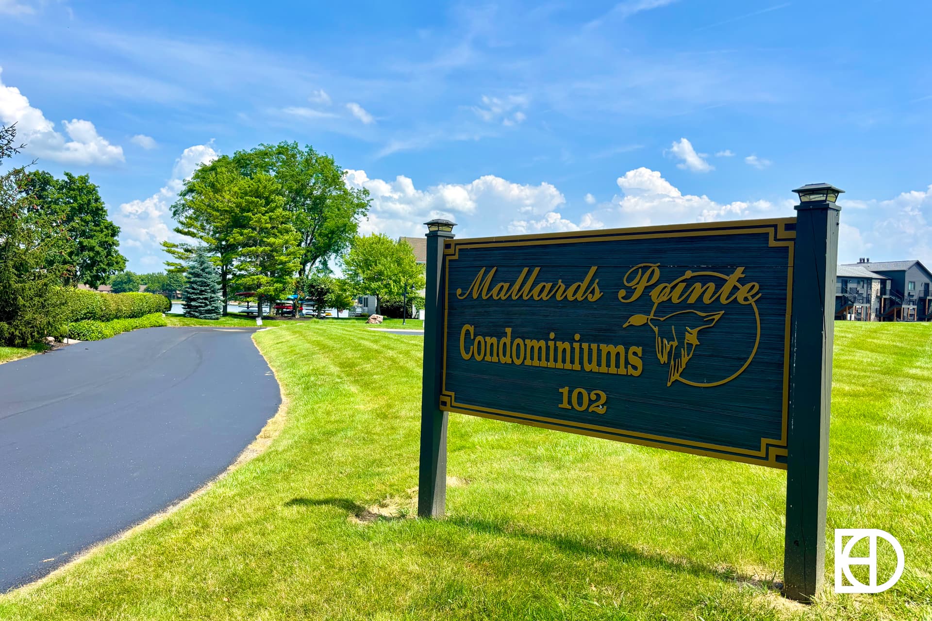 A wooden sign reading Mallards Pointe Condominiums 102 stands on a green lawn beside a paved driveway, with trees, grass, and condos visible under a blue sky with clouds.