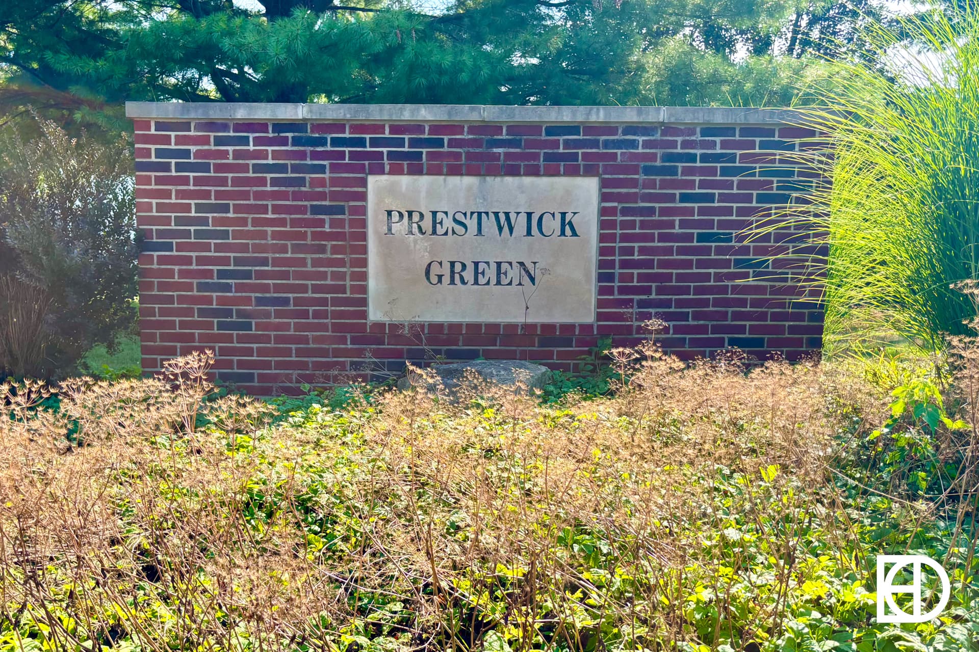 A brick sign with a stone plaque that reads PRESTWICK GREEN stands behind tall grass and greenery, with trees and sunlight in the background. A white KD logo appears in the bottom right corner.