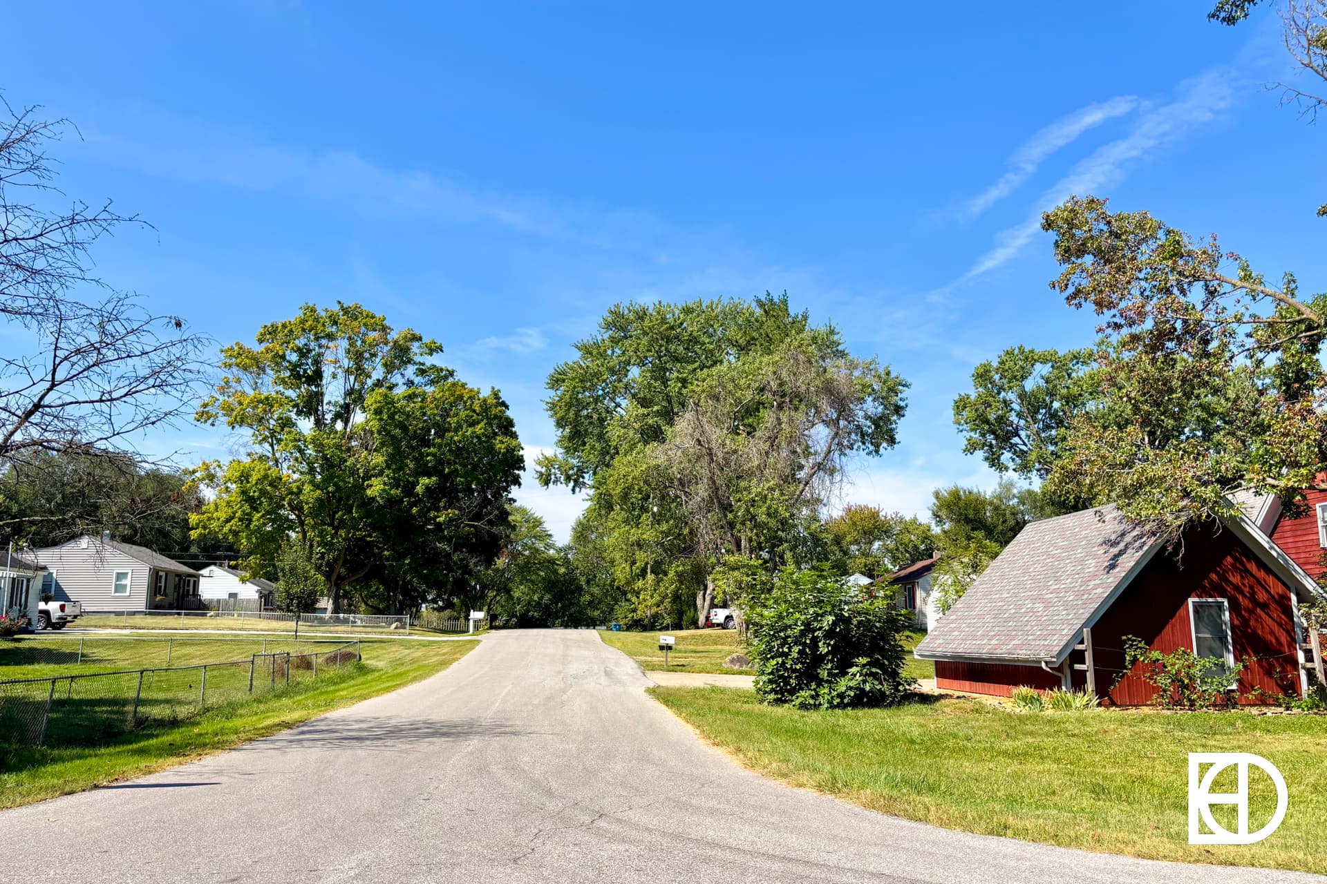 A quiet residential street curves through a neighborhood with green trees, grassy lawns, and small houses under a bright blue sky.