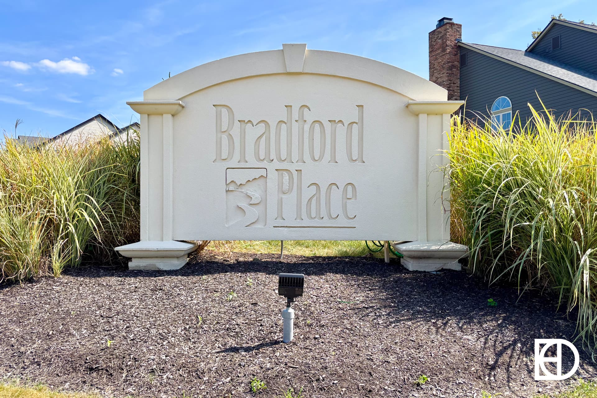A large white sign with raised lettering reads Bradford Place, set among tall grass and mulch in front of residential houses under a blue sky.