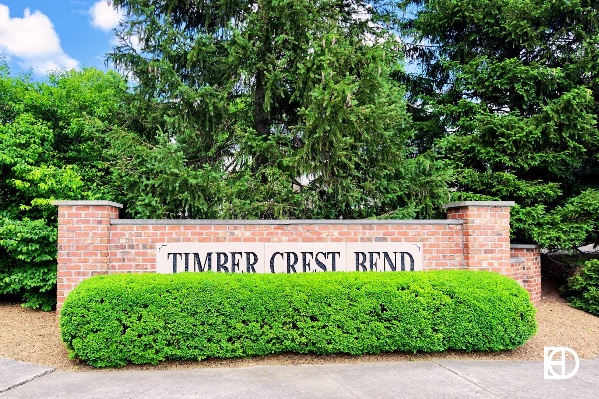 Entrance sign to Timber Crest Bend with brick half wall, trees behind and shrubs in front.