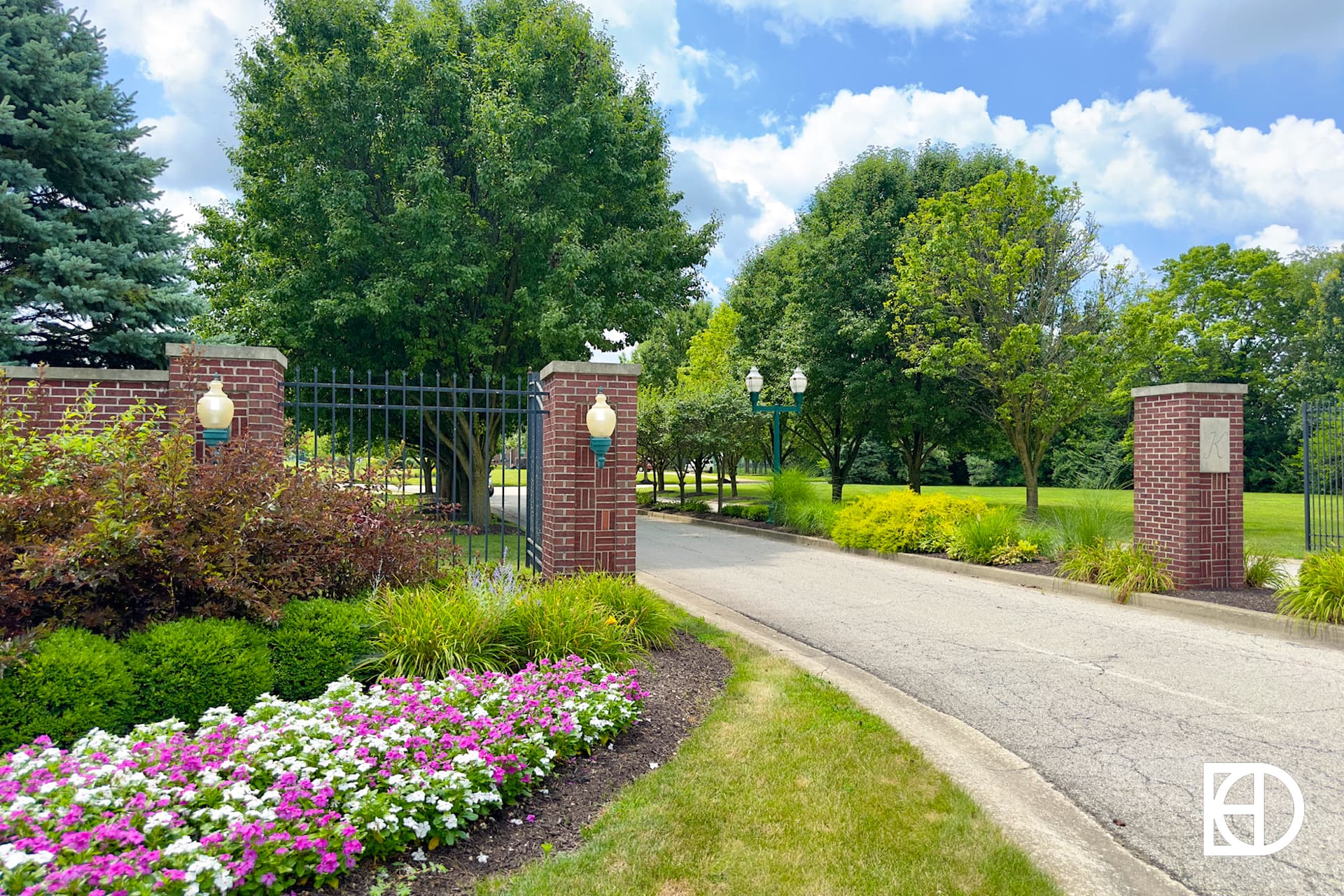 A gated entrance with brick pillars and black iron fencing, surrounded by green trees, manicured grass, and colorful flowerbeds under a partly cloudy sky. A white logo is in the bottom right corner.
