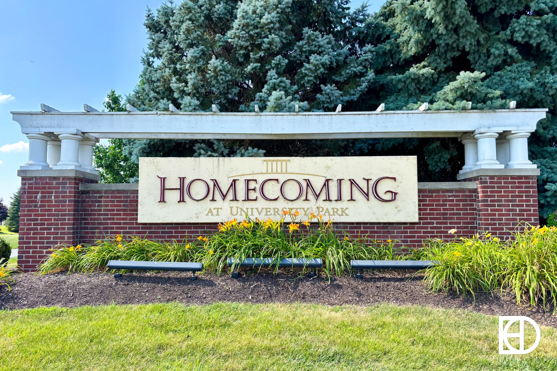 A brick sign with columns reads Homecoming at University Park, surrounded by green grass, orange flowers, and pine trees. The sky is blue and sunny.