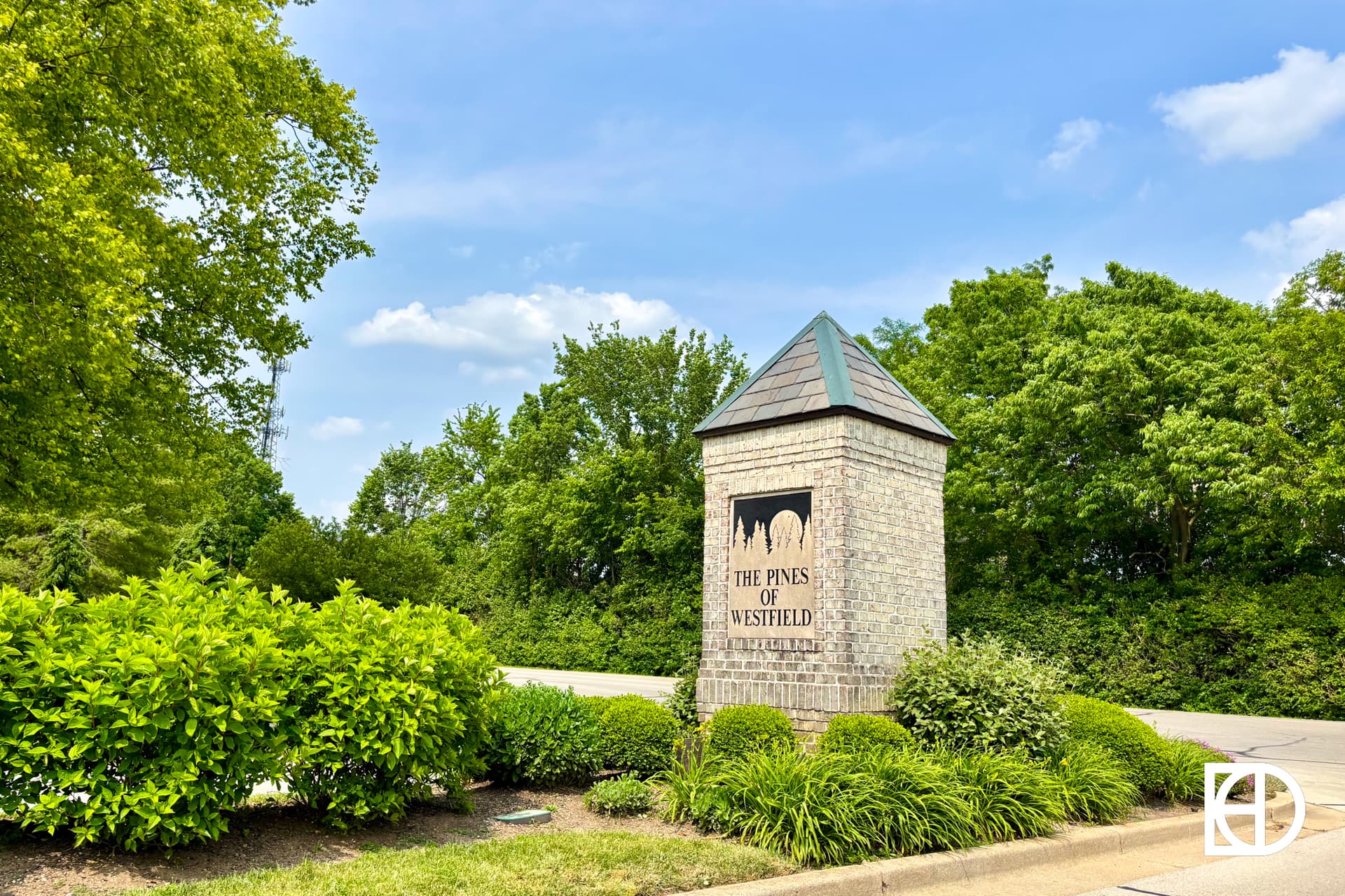A brick monument sign for The Pines of Westfield neighborhood stands surrounded by green shrubs and trees under a partly cloudy sky.