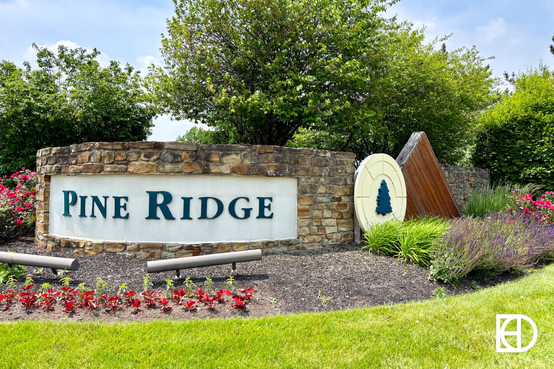 A stone entrance sign reads Pine Ridge, surrounded by landscaped bushes, flowers, and a decorative emblem featuring a pine tree.
