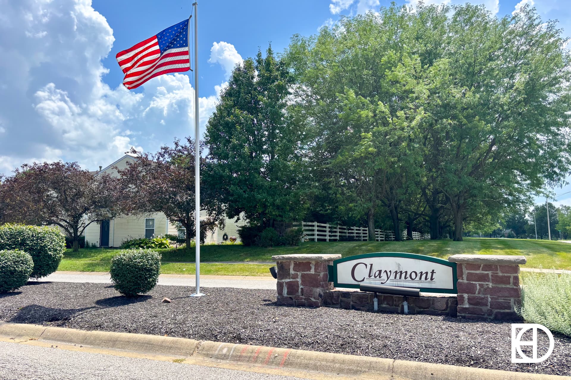 A stone sign reading Claymont stands by a roadside, bordered by brick pillars and mulch landscaping. An American flag flies on a pole nearby, with trees and a house visible in the background under a blue sky.