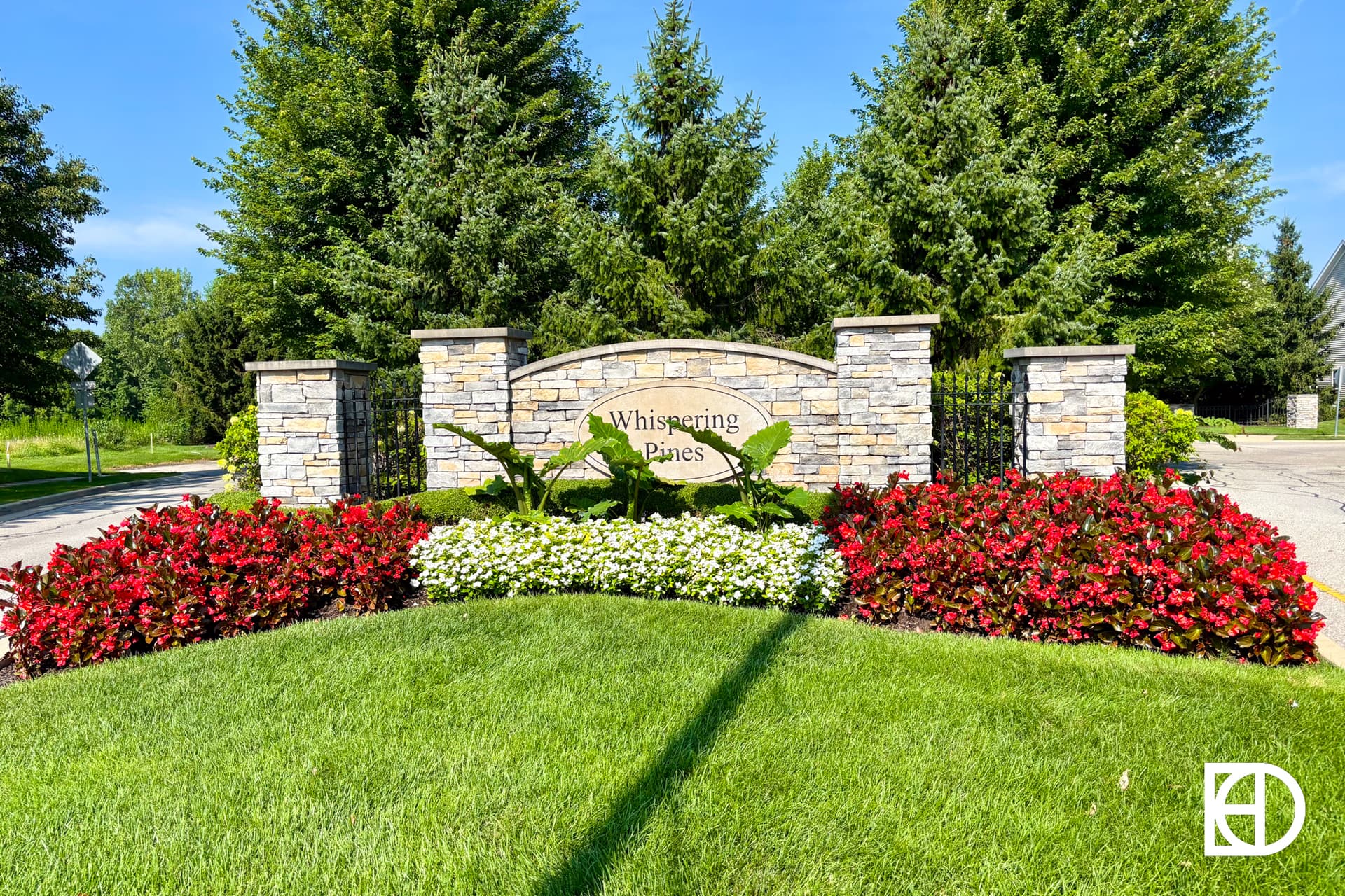 A stone entrance sign reading Whispering Pines is surrounded by vibrant red and white flowers, lush green grass, and tall evergreen trees in the background.
