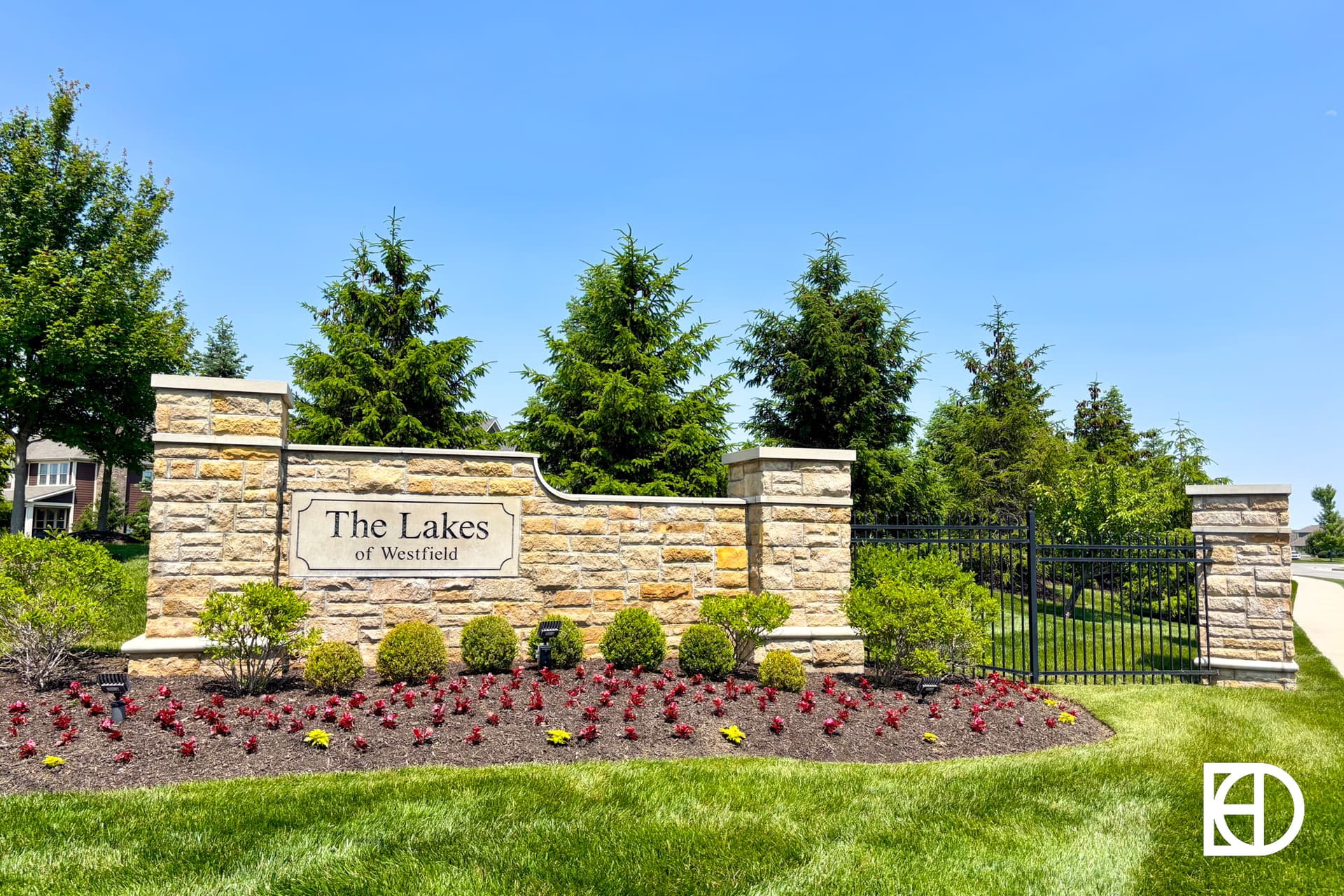 Stone entrance sign reading The Lakes of Westfield, surrounded by landscaped flower beds, green grass, and trees under a clear blue sky.