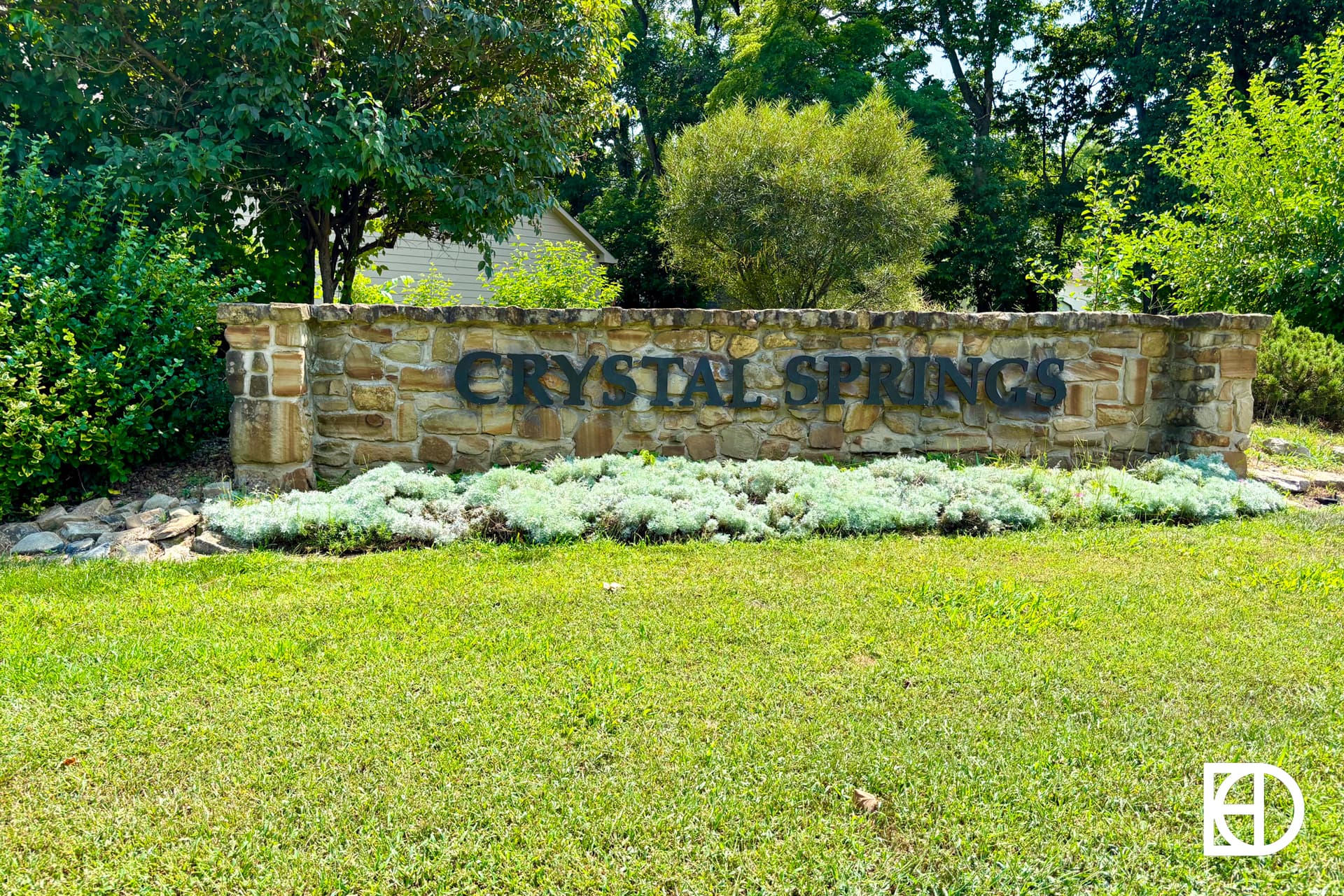 A brick sign with “Ashley Oakes” in black letters is surrounded by bushes and trees. A house with a sloped roof is partially visible behind the sign under a partly cloudy sky.
