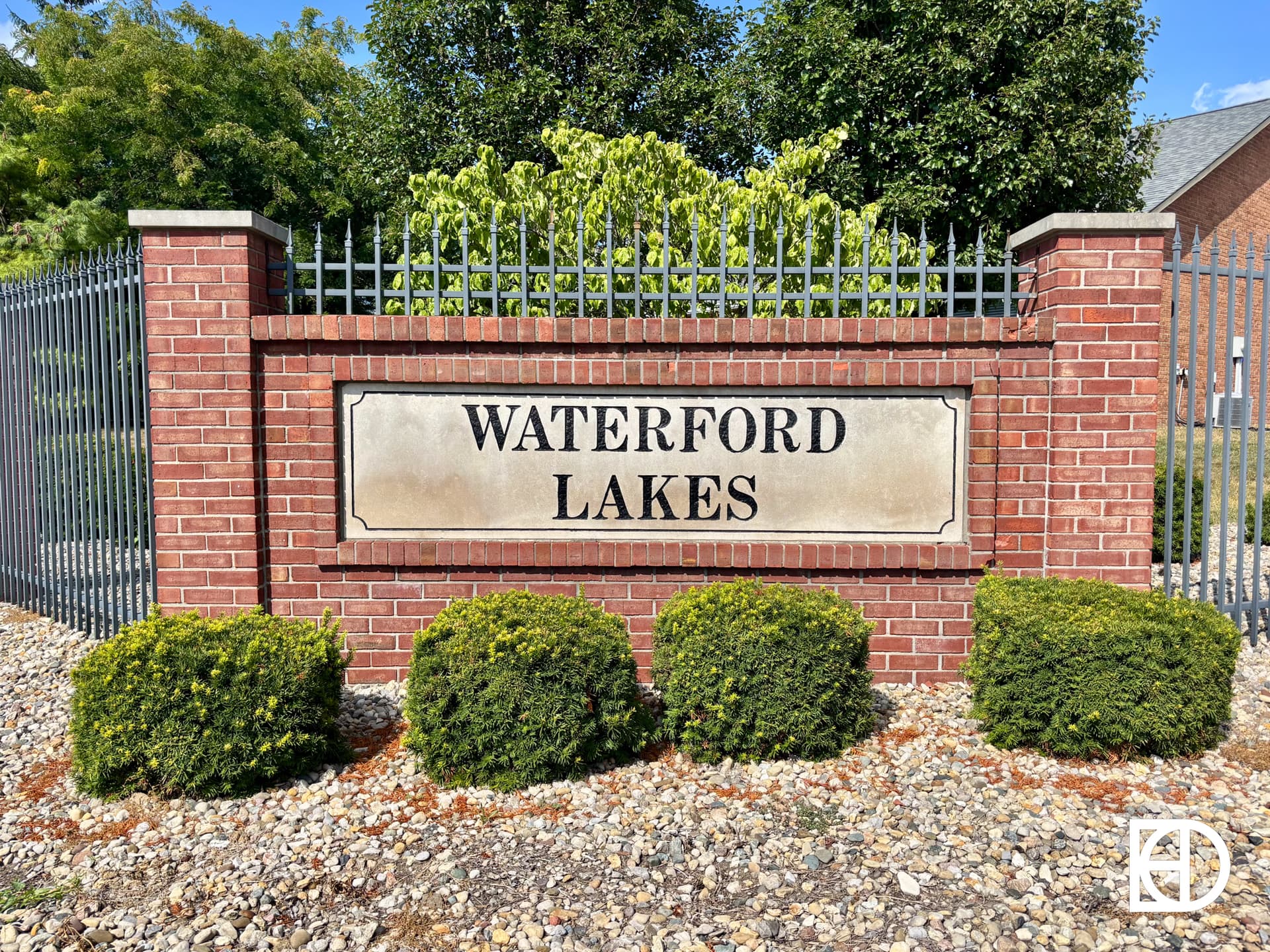 A brick sign with the text Waterford Lakes is displayed between metal fence posts, with bushes in front and trees in the background.