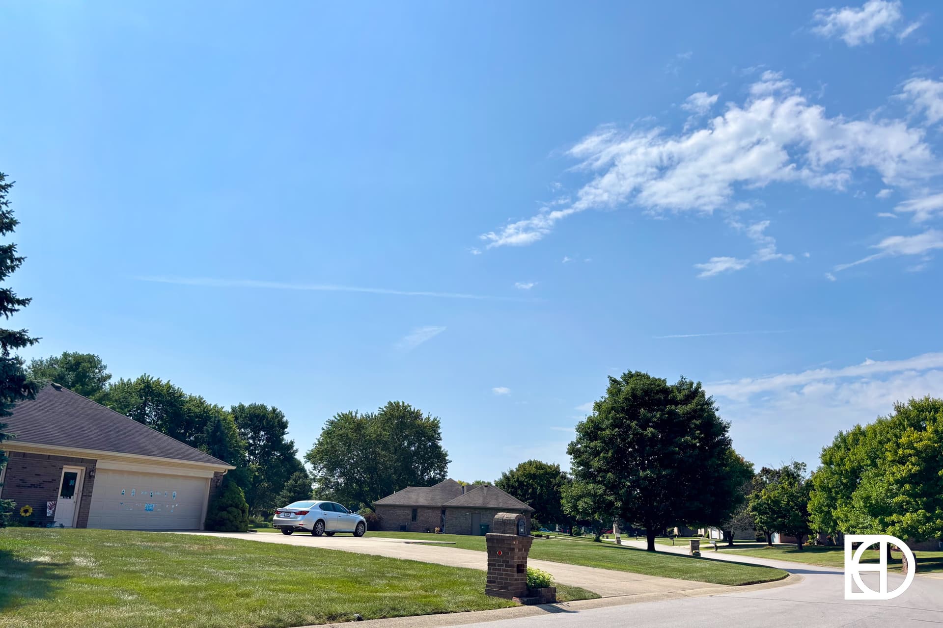 A suburban neighborhood with green lawns, trees, a parked white car in a driveway, and houses under a clear blue sky with a few clouds. A brick mailbox is visible in the foreground.