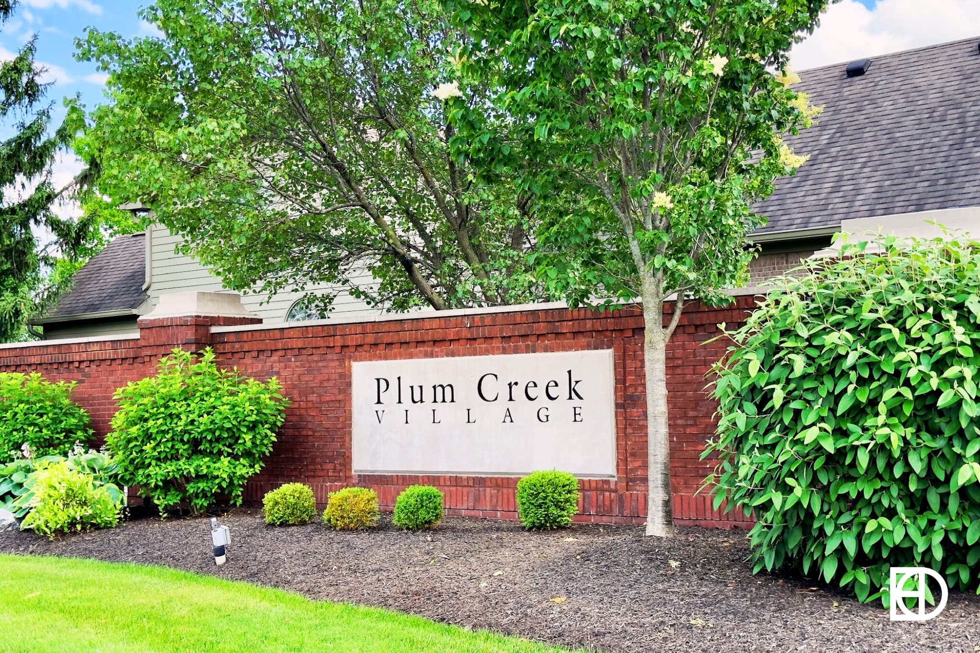 Brick and stone entrance sign for Plum Creek Village with shrubs around and trees behind.