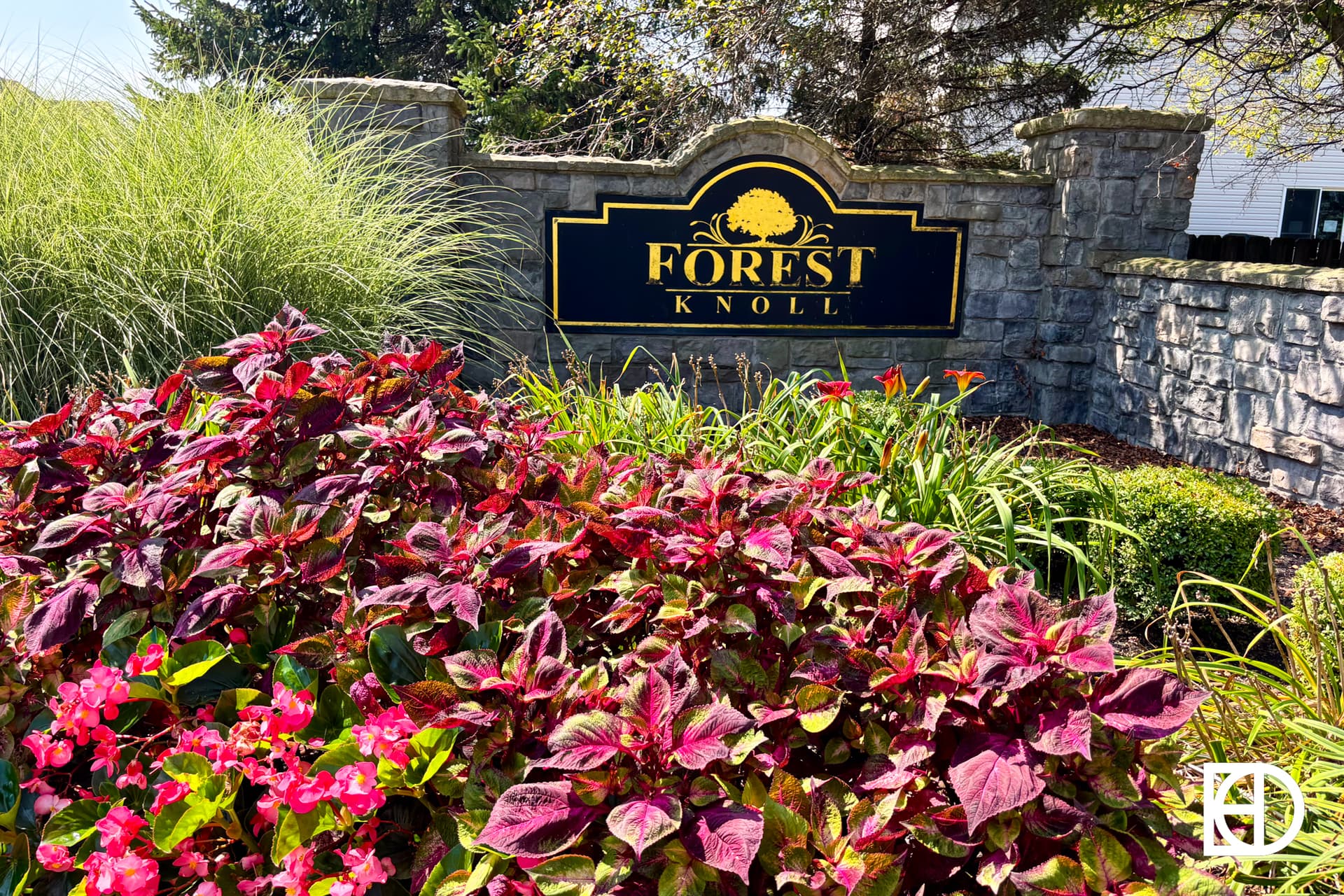 A stone entrance sign reading Forest Knoll is surrounded by vibrant red and pink flowers, green shrubs, and tall ornamental grasses under bright sunlight.