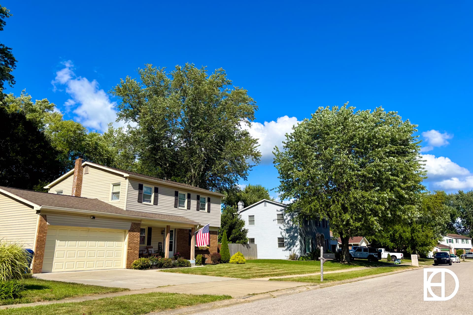 A suburban street with two-story houses, trees, a bright blue sky, and a few clouds. An American flag hangs by the garage of the nearest house. Several cars are parked along the street.
