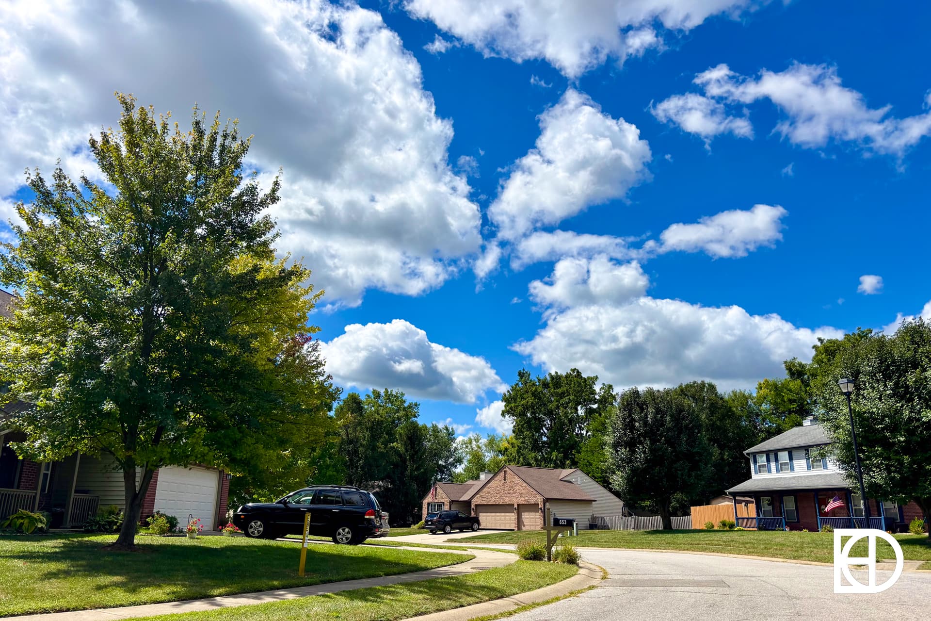 A suburban neighborhood street on a sunny day, with a few parked cars, green lawns, trees, and houses under a blue sky filled with fluffy white clouds.