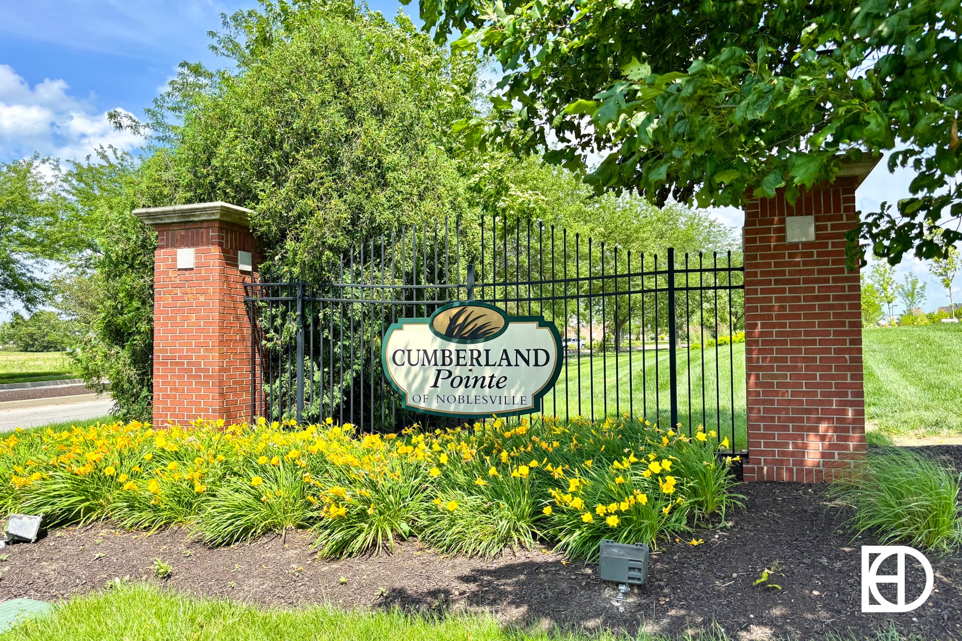 Entrance sign for Cumberland Pointe of Noblesville, set on a black metal fence between two red brick pillars, surrounded by yellow flowers and green shrubs, with trees and grass in the background.