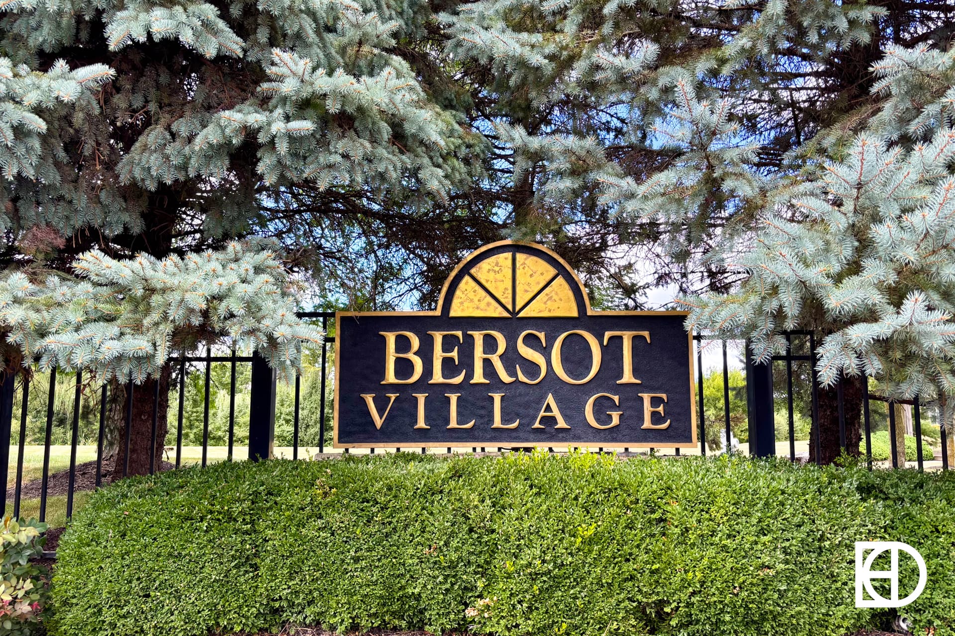 A black and gold sign reading Bersot Village is surrounded by neatly trimmed bushes and tall evergreen trees, with a black metal fence in the background.