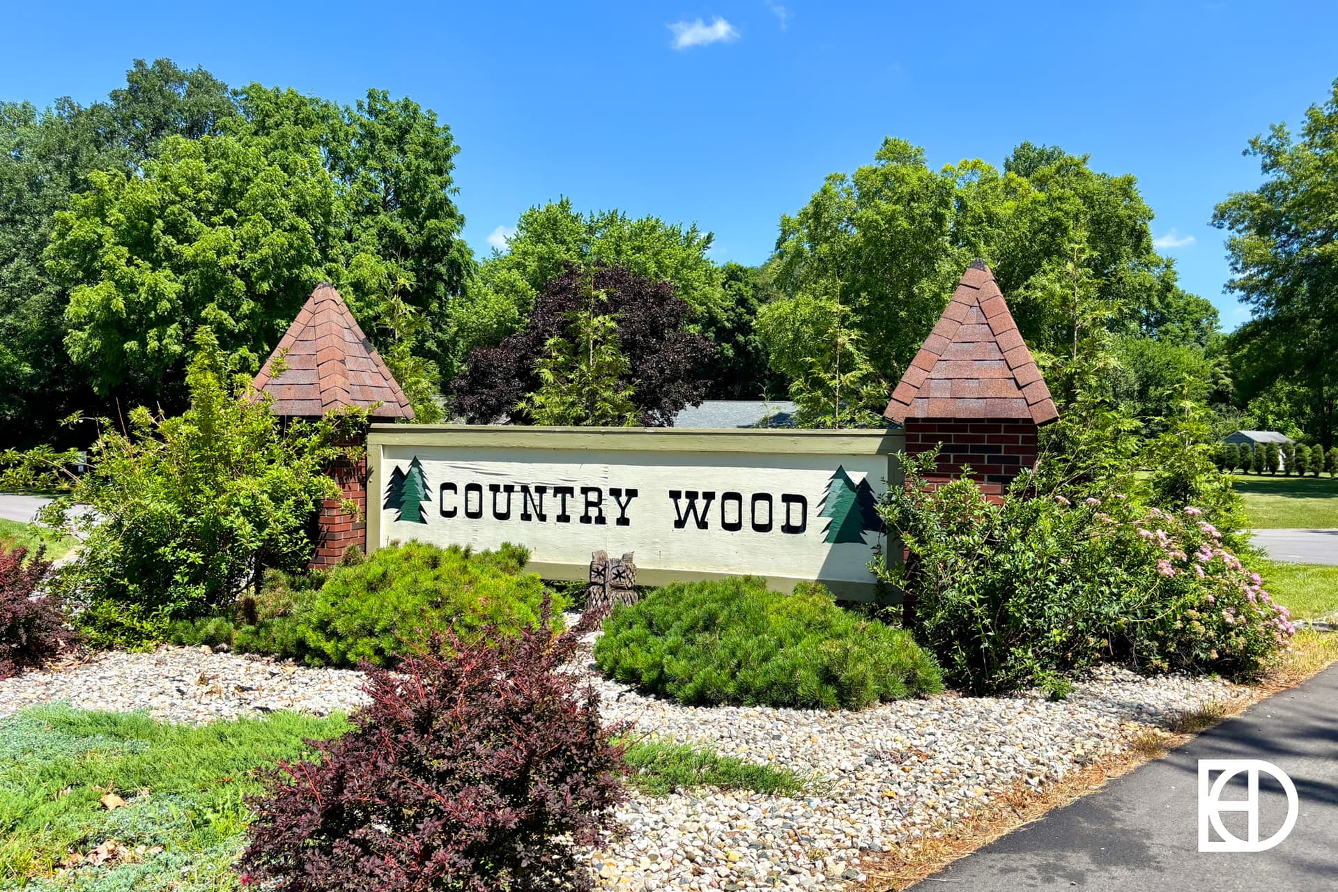 A brick sign with two peaked columns reads Country Wood and features green pine tree graphics, surrounded by lush bushes and trees under a clear blue sky