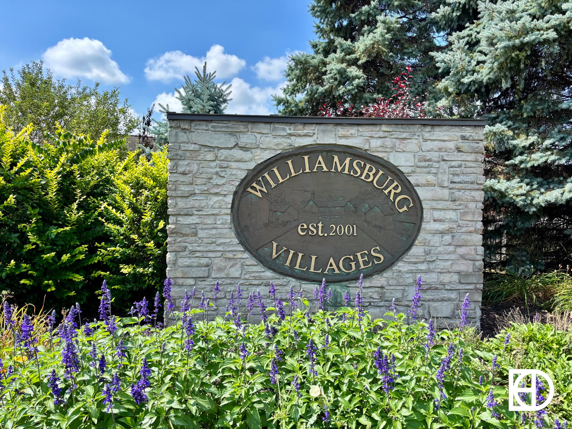 A stone sign with an oval plaque reads Williamsburg Villages est. 2001, surrounded by green shrubs, purple flowers, and pine trees under a blue sky with clouds.
