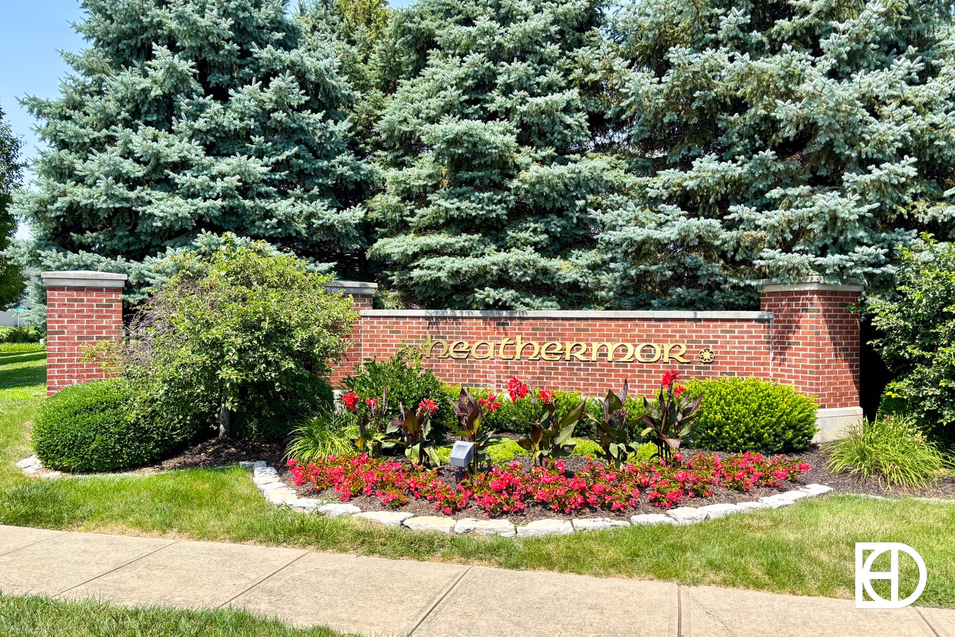 A brick sign with the word Heathermoor in gold letters stands behind a flower bed with red and pink flowers, green shrubs, and tall evergreen trees in the background.
