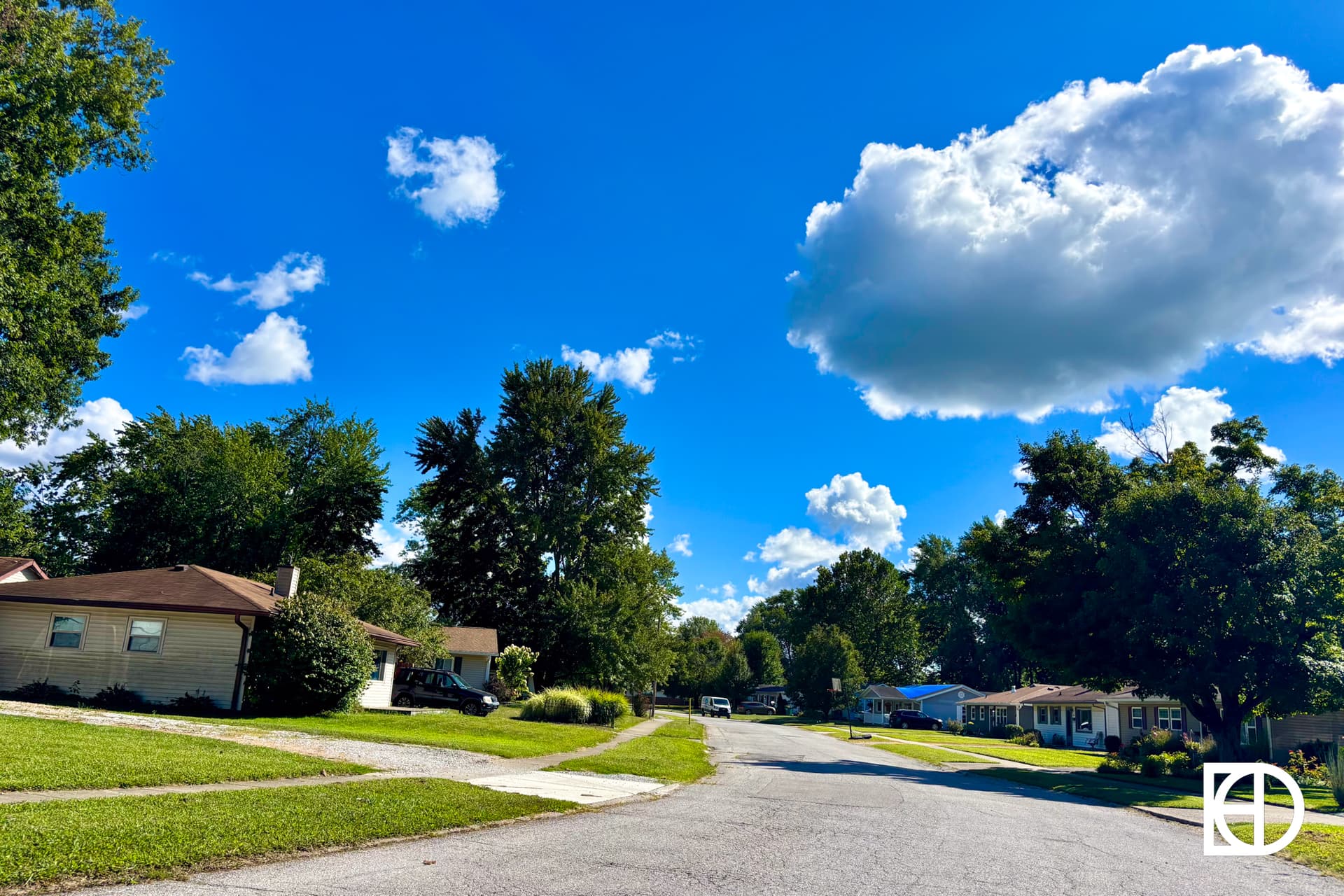 A quiet suburban street with houses and green lawns under a bright blue sky with scattered white clouds. Trees line both sides of the street, and sunlight casts shadows on the road and grass.