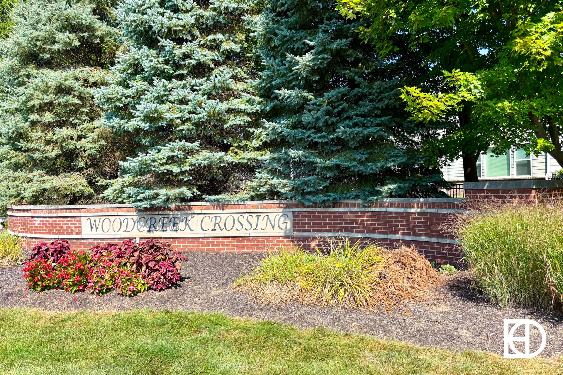 A brick sign reading Woodcreek Crossing stands in front of evergreen trees and shrubs, with landscaped greenery and a lawn in the foreground.