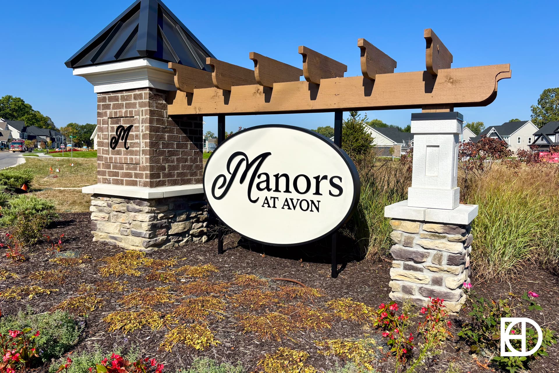 A landscaped entrance sign with Manors at Avon written in black script on a large white oval, supported by stone columns and a wooden pergola, with houses and flowers in the background.
