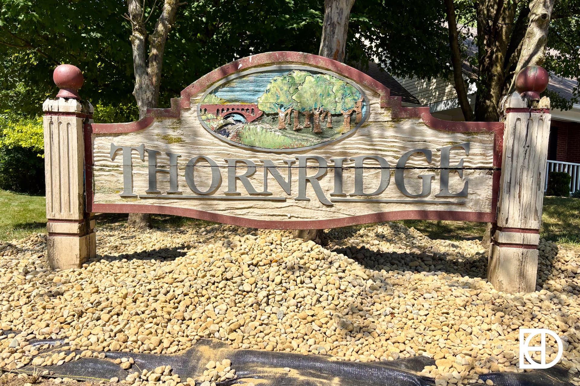 A wooden sign reads “Thornridge” with a carved landscape scene above the text, featuring trees and a bridge. The sign is set on gravel with trees and a house in the background.