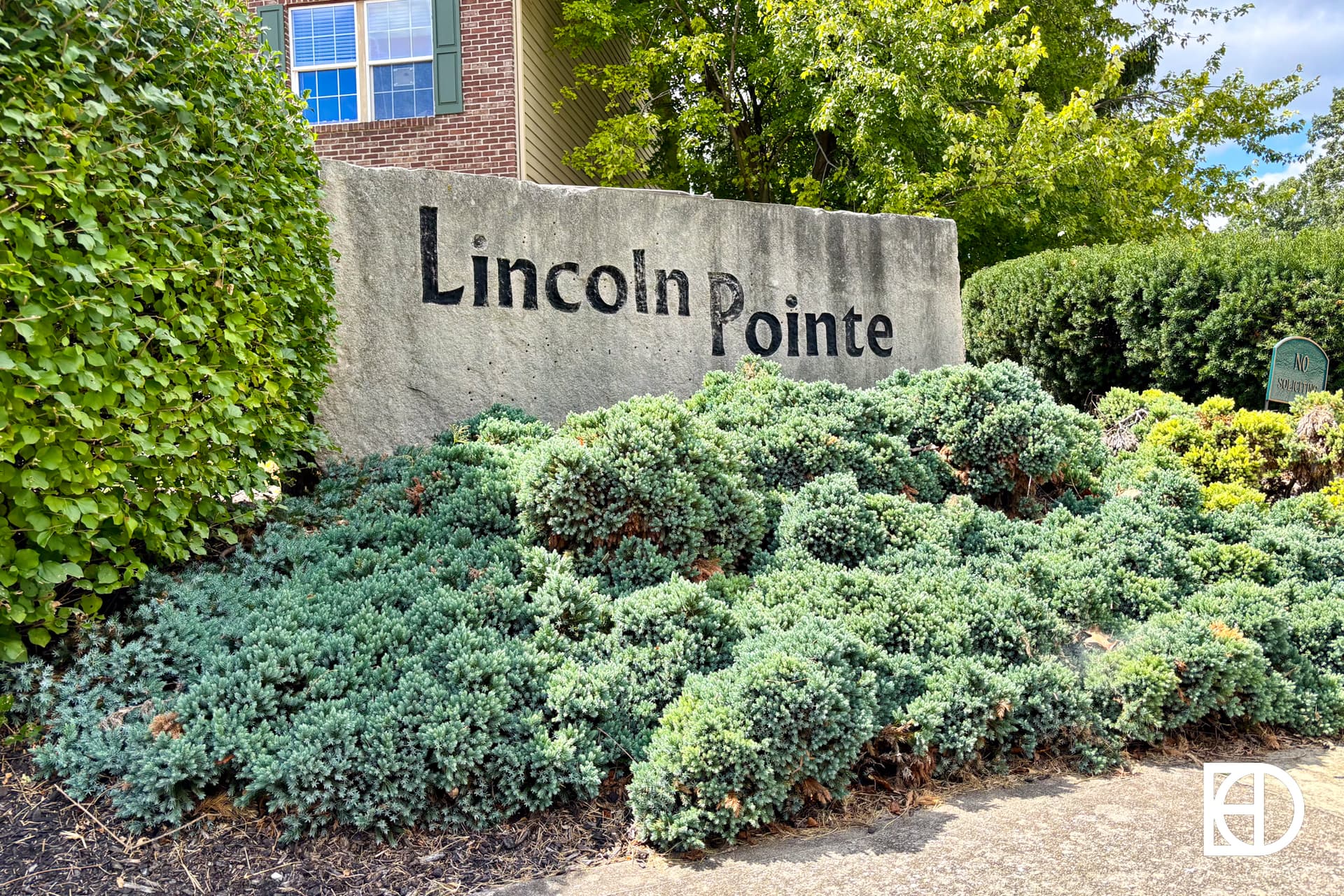 A large stone sign with Lincoln Pointe written on it sits behind green bushes and landscaping, with a brick building and trees visible in the background.