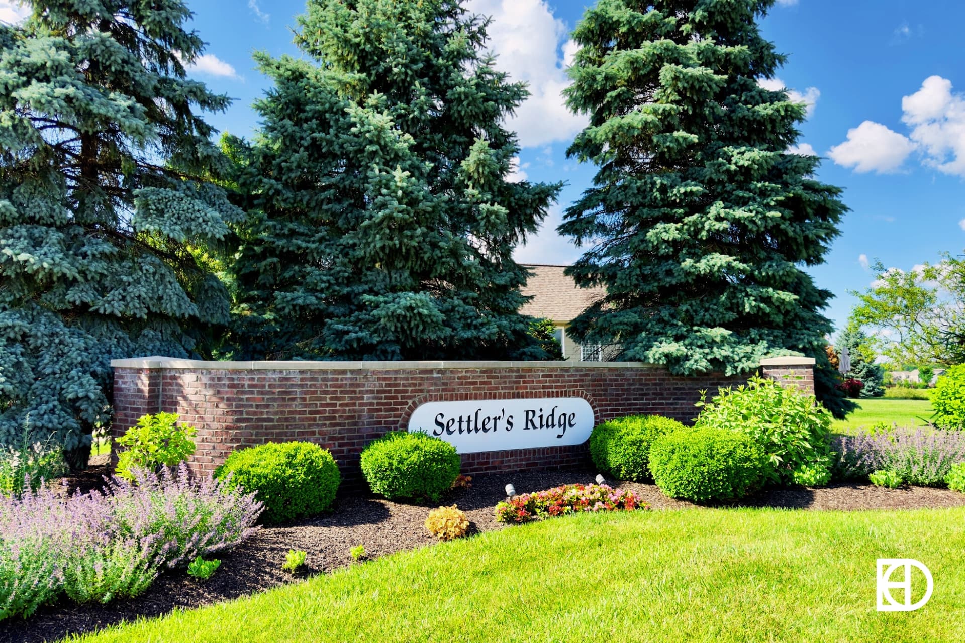 Brick half wall entrance sign to Settler's Ridge with evergreen trees behind and shrubs and flowers surrounding.