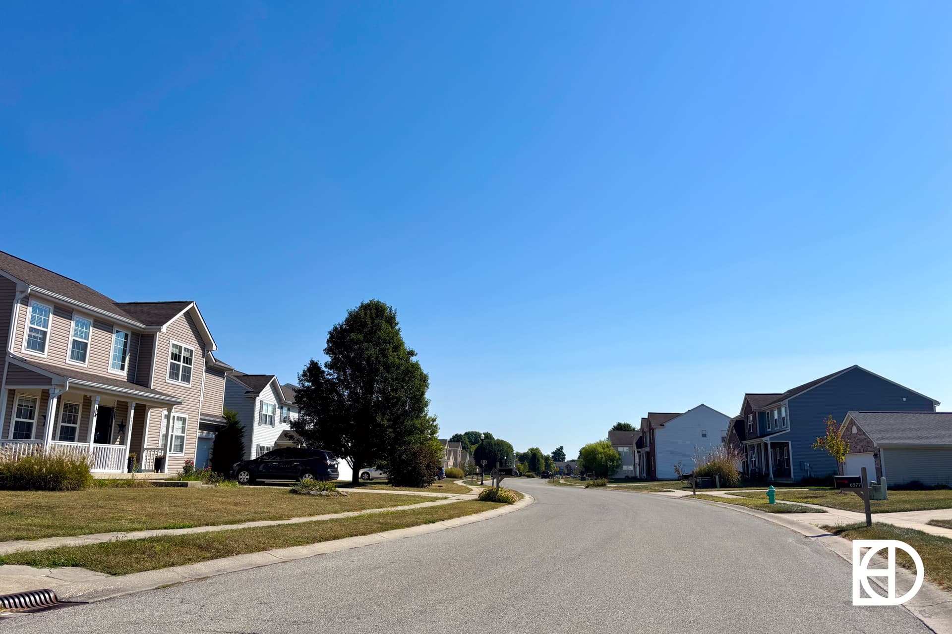 A quiet suburban street curves through a neighborhood with two-story houses, green lawns, trees, and a bright blue sky overhead.