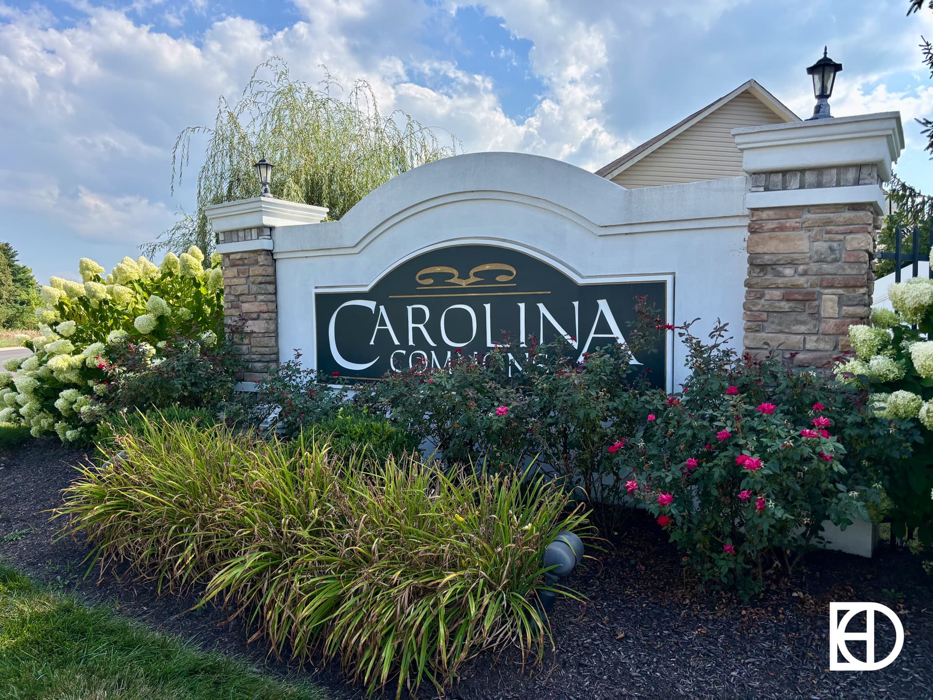 A landscaped entrance sign for Carolina Commons, surrounded by flowering shrubs and ornamental grasses, with a blue sky and part of a house in the background. The KD logo appears in the lower right corner.