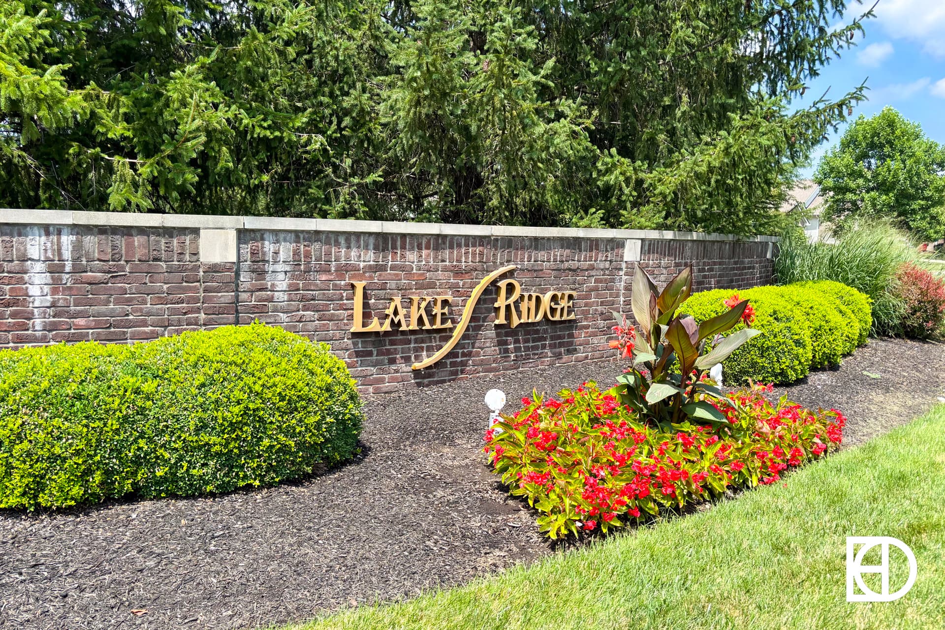 A brick sign reading Lake Ridge is surrounded by neatly trimmed bushes, red flowers, and green grass, with tall pine trees in the background. A white KD logo appears in the bottom right corner.