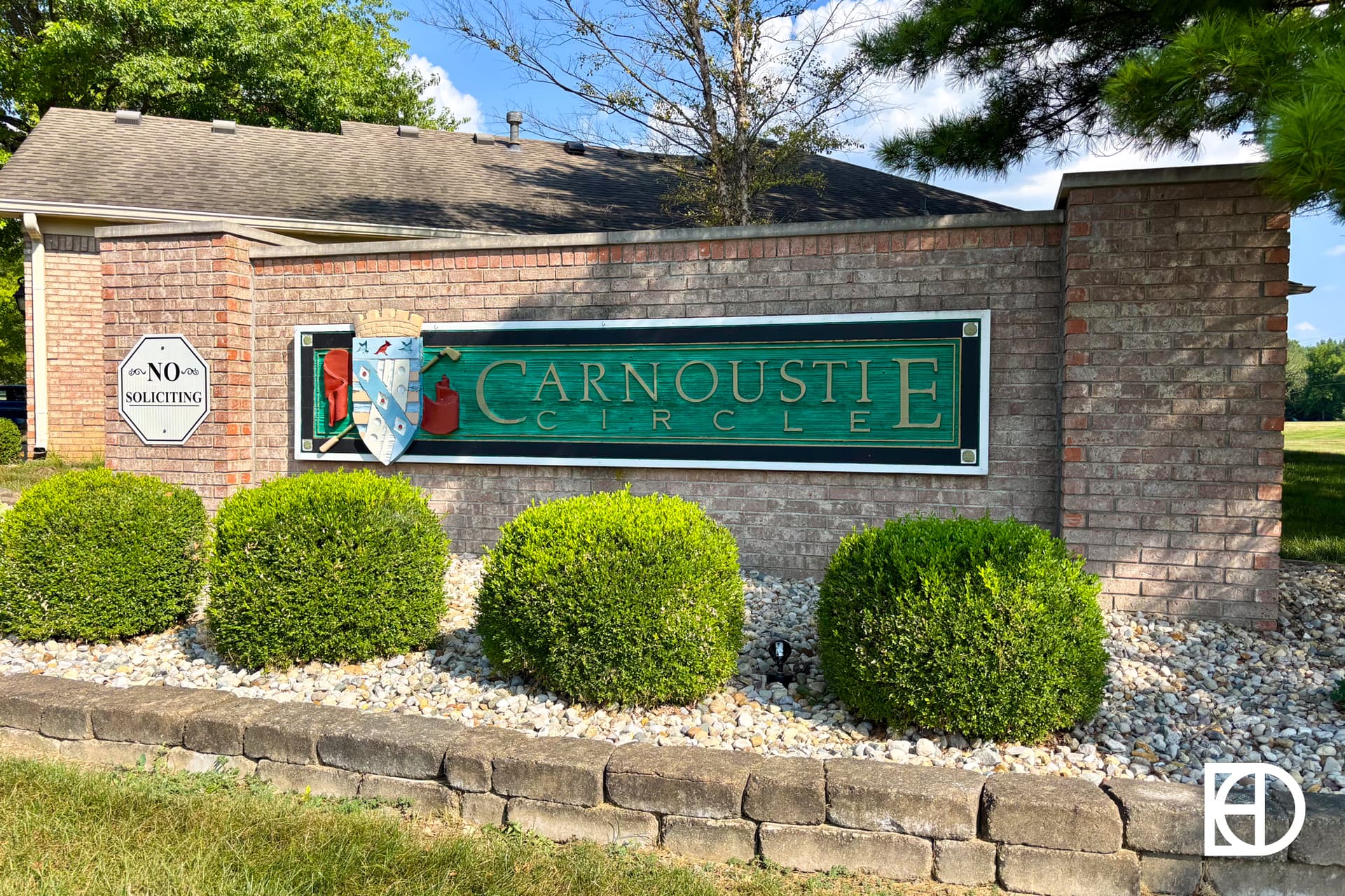 A brick entrance sign for Carnoustie Circle features a green plaque with a crest, surrounded by neatly trimmed bushes and a No Soliciting sign on the left. The KD logo is in the bottom right corner.