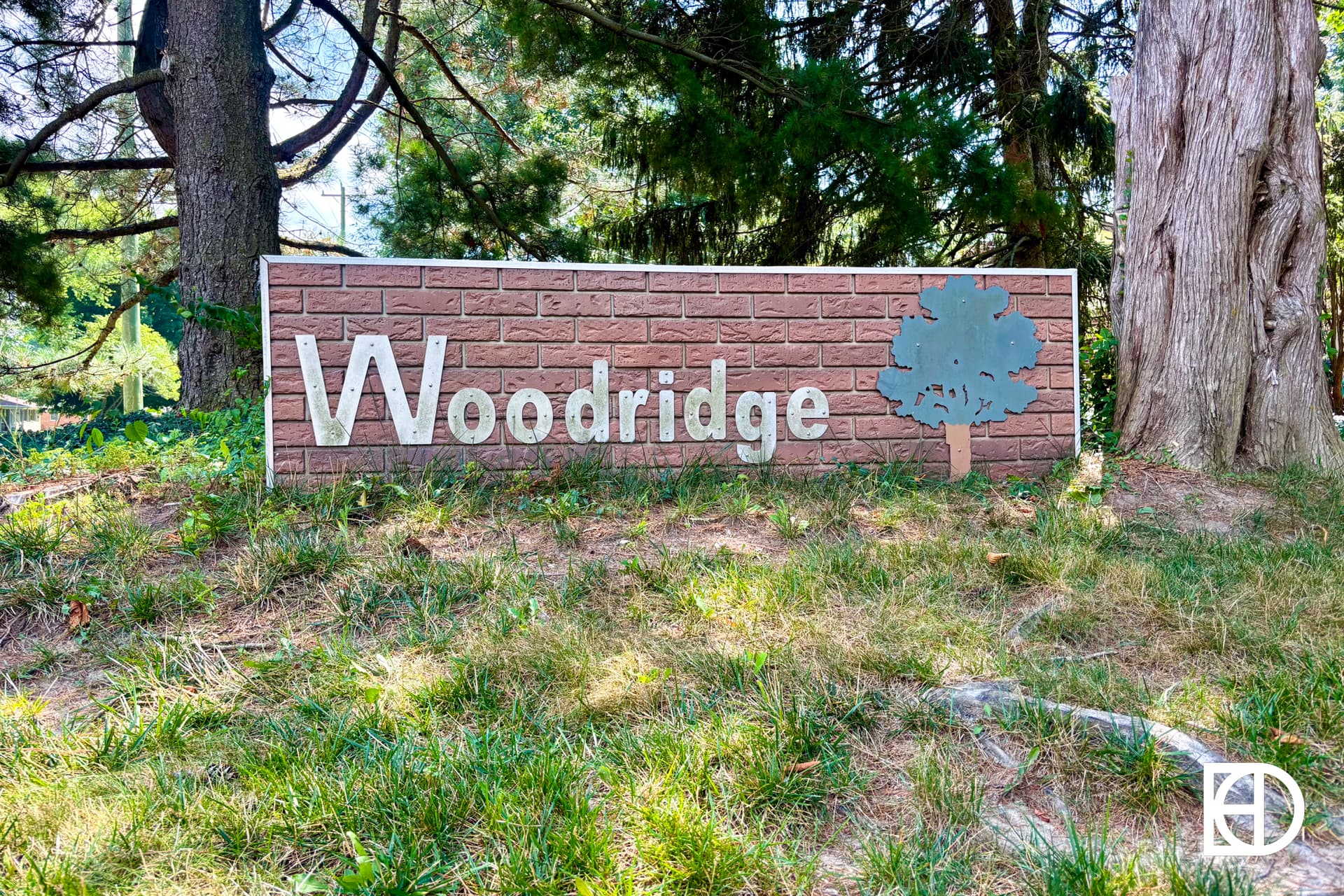 A brick-style sign reading Woodridge with a tree graphic, surrounded by grass and tall trees in a shaded outdoor area.
