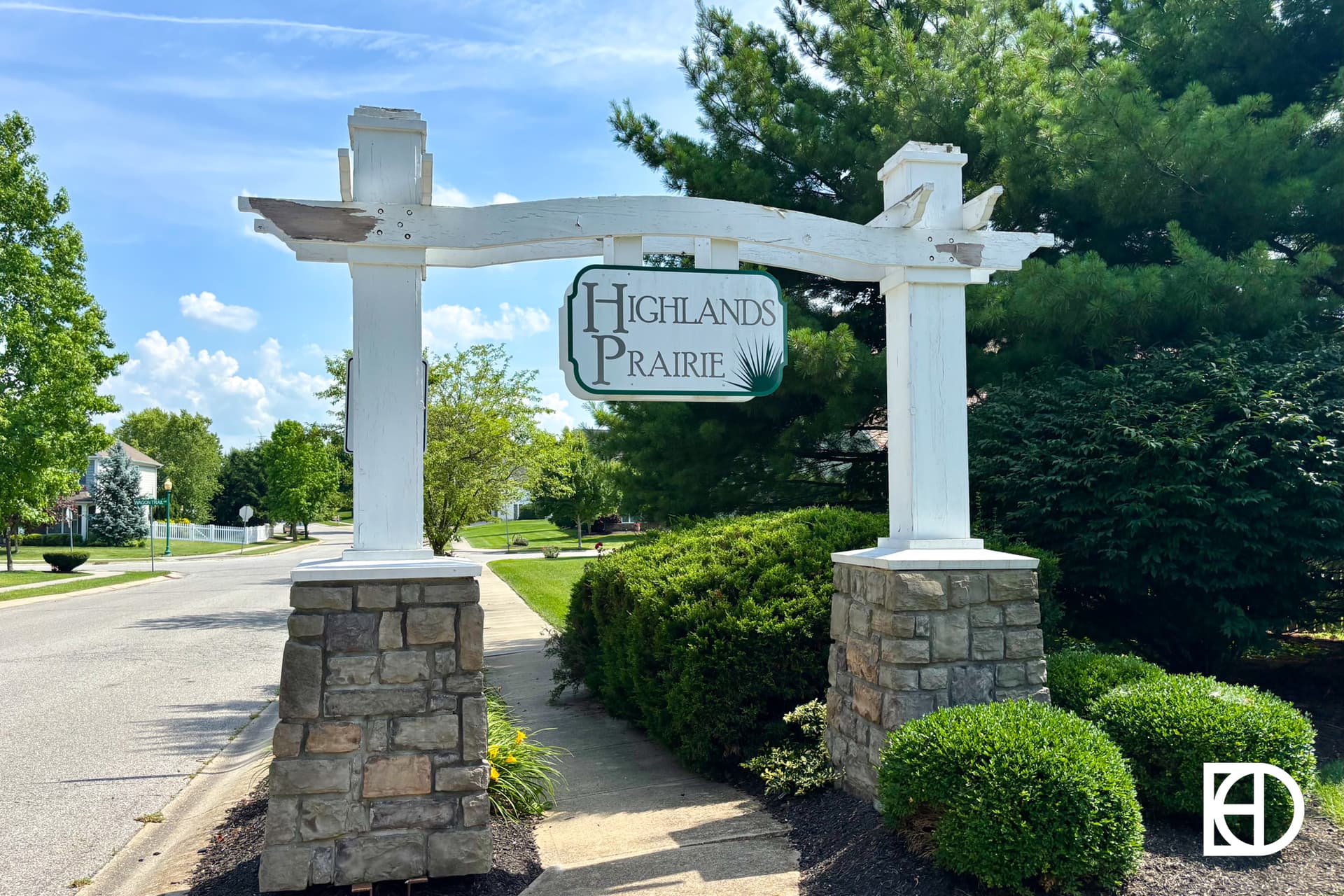 A wooden sign reading Highlands Prairie is mounted on a white frame with stone columns, standing by a sidewalk surrounded by green bushes and trees on a sunny day in a suburban neighborhood.