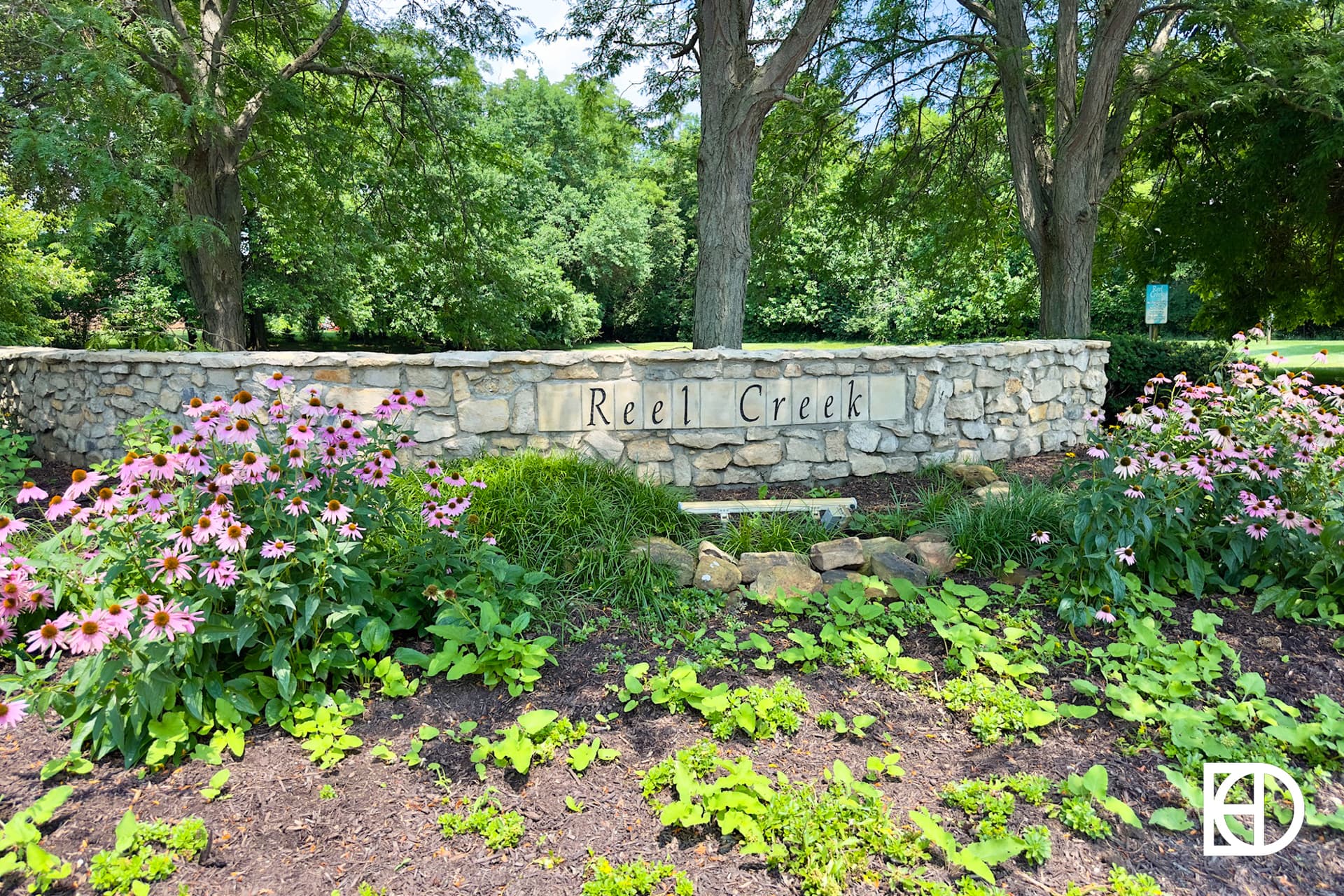 A stone wall with the words Reel Creek is surrounded by pink coneflowers, green plants, and trees on a sunny day. A white ED logo is in the bottom right corner.