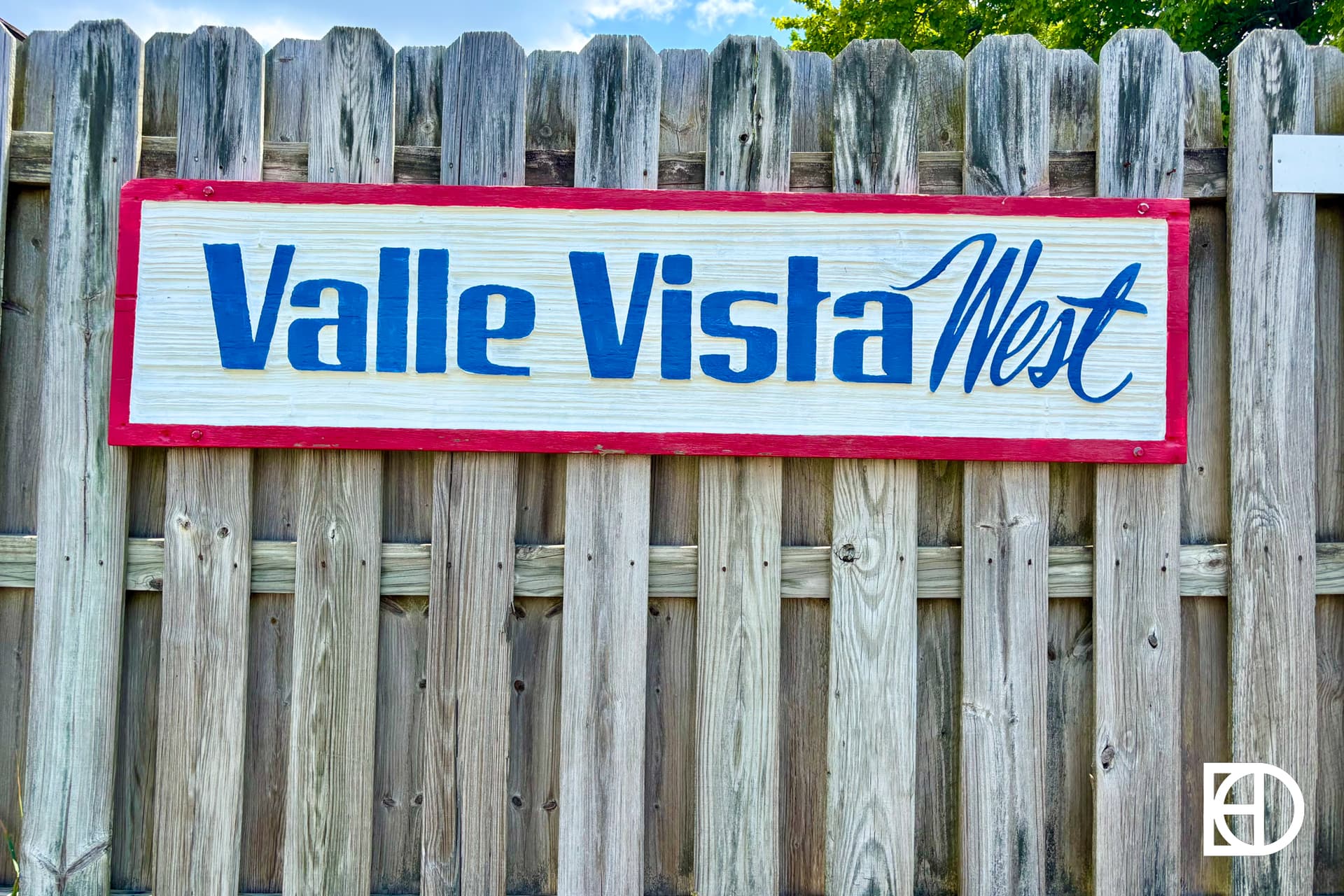 A suburban street on a sunny day with houses, green lawns, trees, and a bright blue sky filled with scattered clouds. The KD logo appears in the bottom right corner.