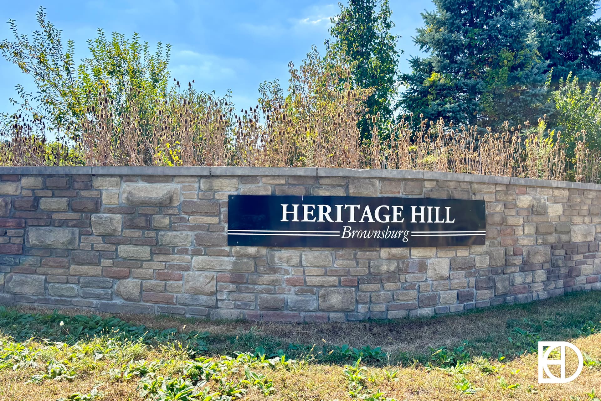 Stone wall with a black sign that reads HERITAGE HILL Brownsburg in white letters, surrounded by plants and trees under a clear blue sky.