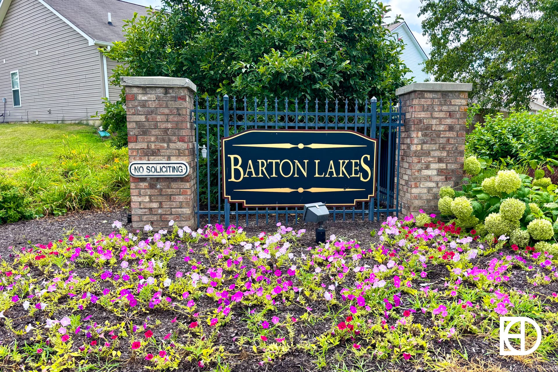 A brick and metal sign reading Barton Lakes stands behind colorful blooming flowers, with a No Soliciting sign attached to the fence and houses and greenery visible in the background.